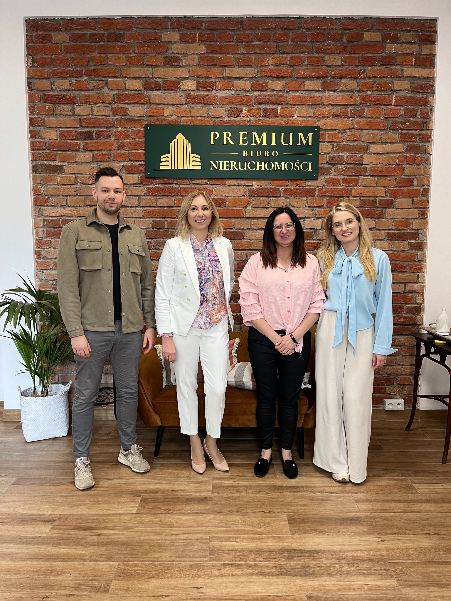 Grupa czterech osób (dwóch mężczyzn i dwie kobiety) stojących przed ceglaną ścianą z szyldem 'PREMIUM BIURO NIERUCHOMOŚCI', z widoczną rośliną w doniczce i fragmentem sofy w tle.