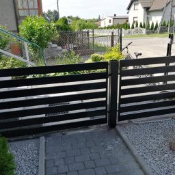 ParvisWelder Michał Gawarkiewicz - Nowoczesna, pozioma brama wjazdowa z czarnych, metalowych profili, widoczna od strony posesji, z brukowanym podjazdem i ozdobnym kamieniem na żwirze.