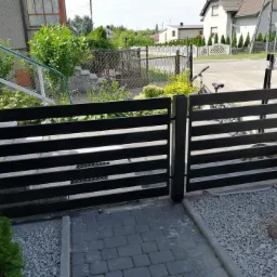 Nowoczesna, pozioma brama wjazdowa z czarnych, metalowych profili, widoczna od strony posesji, z brukowanym podjazdem i ozdobnym kamieniem na żwirze.