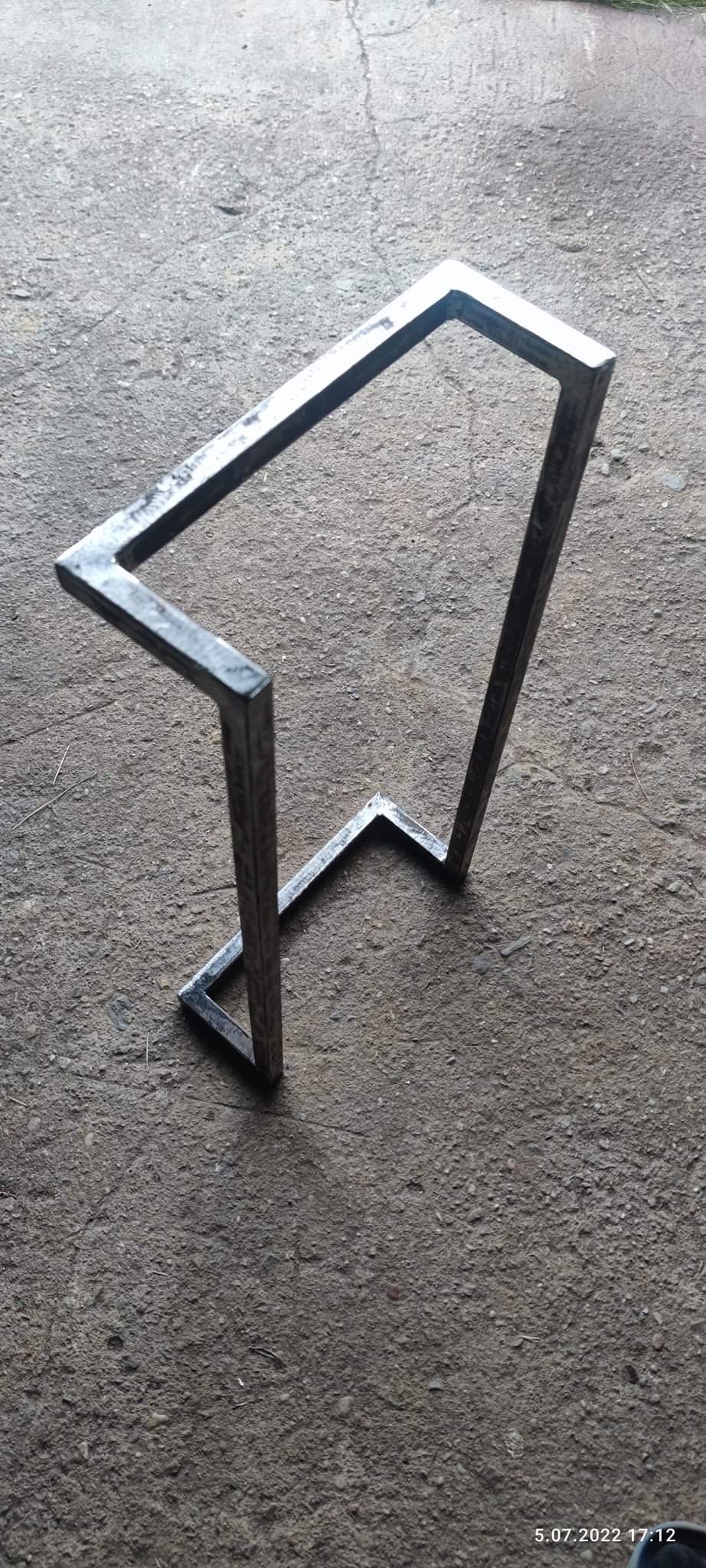 Surowy, spawany metalowy stelaż o kwadratowym przekroju, stojący na szarej, betonowej powierzchni. Widoczne ślady spawania i niedoskonałości wykończenia.