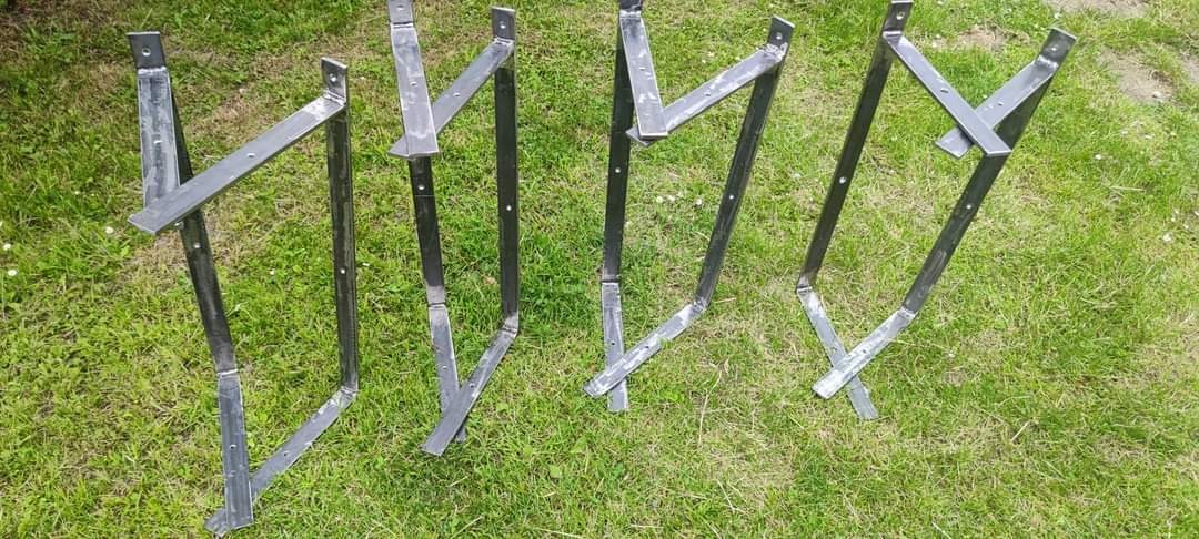 Surowe, spawane, metalowe konstrukcje wspornikowe z otworami na śruby, leżące na trawie w świetle dziennym, prawdopodobnie przygotowane do montażu ogrodzenia lub bramy.