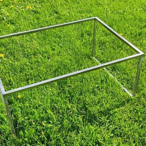 Prosta, spawana konstrukcja z metalowych profili, w kształcie prostopadłościanu, ustawiona na trawie z żółtymi kwiatami.