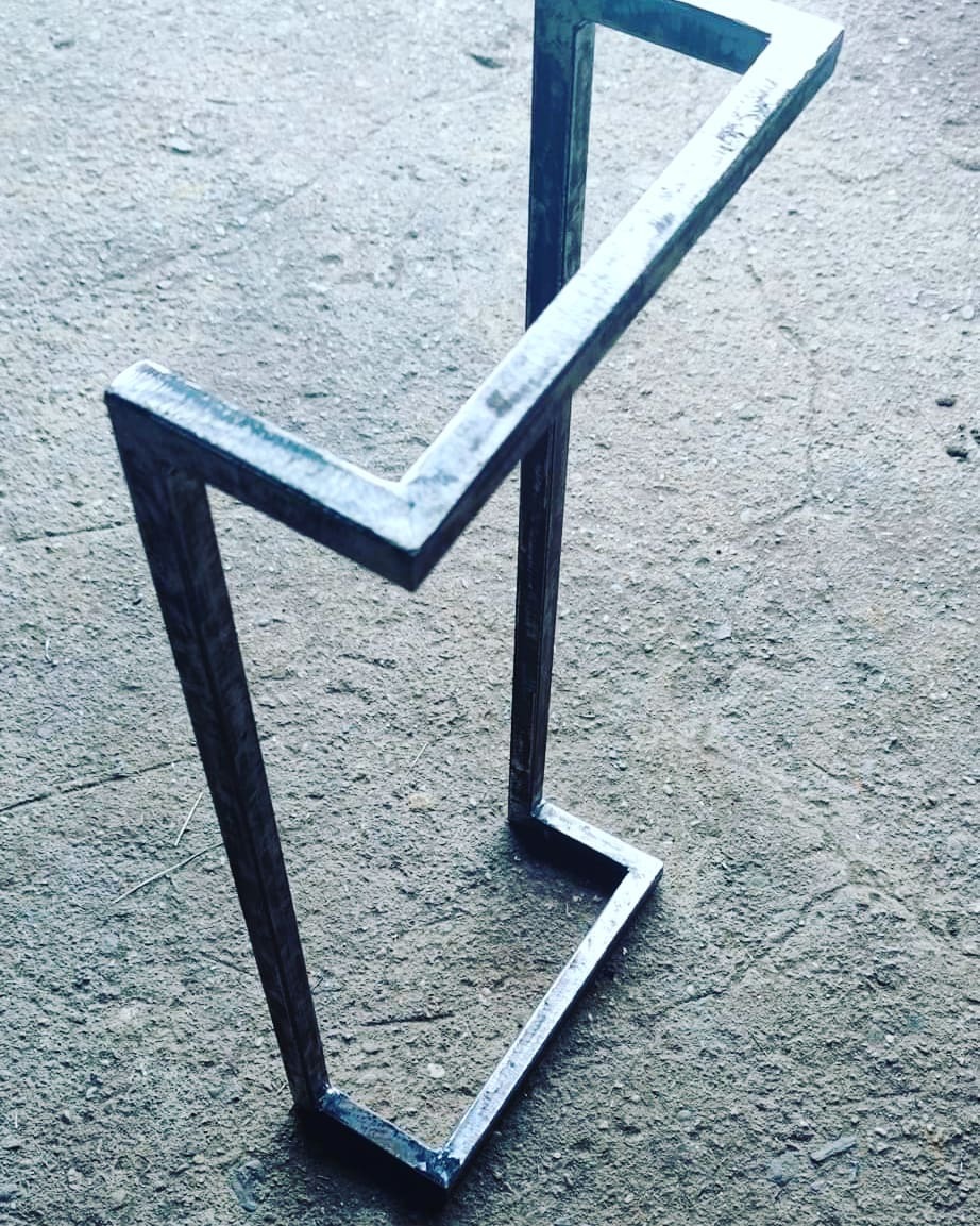 Surowy, geometryczny stojak metalowy z kwadratowych profili, spawany, widoczny na szarym betonowym podłożu.