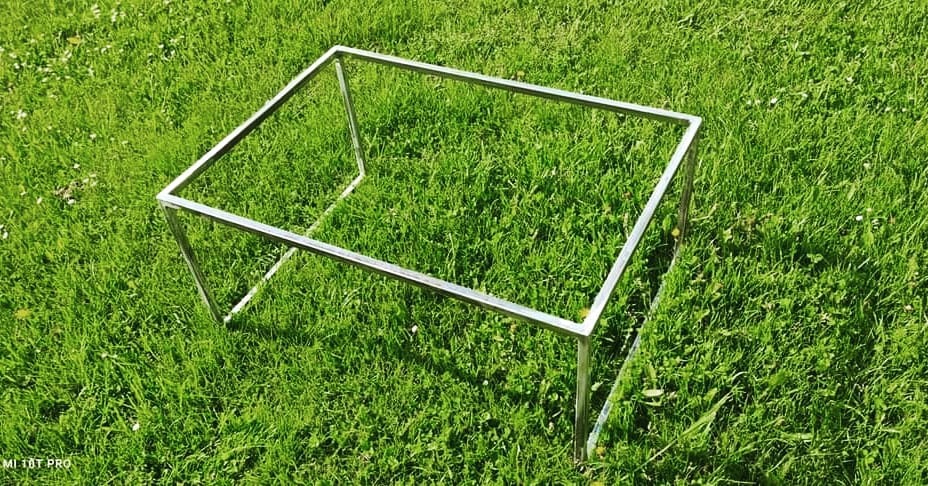 Surowa, spawana konstrukcja z metalowych profili w kształcie prostopadłościanu, umieszczona na trawniku z widocznymi białymi kwiatami.