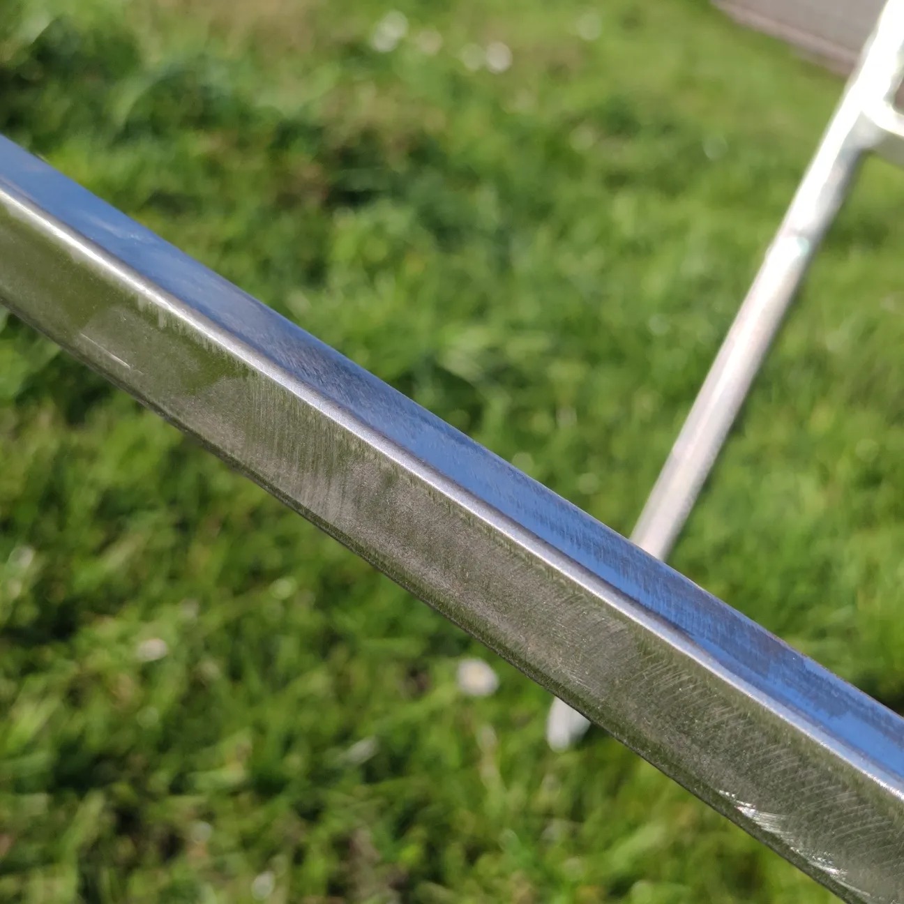 Szlifowany profil metalowy o przekroju kwadratowym, widoczne ślady obróbki, w tle trawa i fragment drabiny.