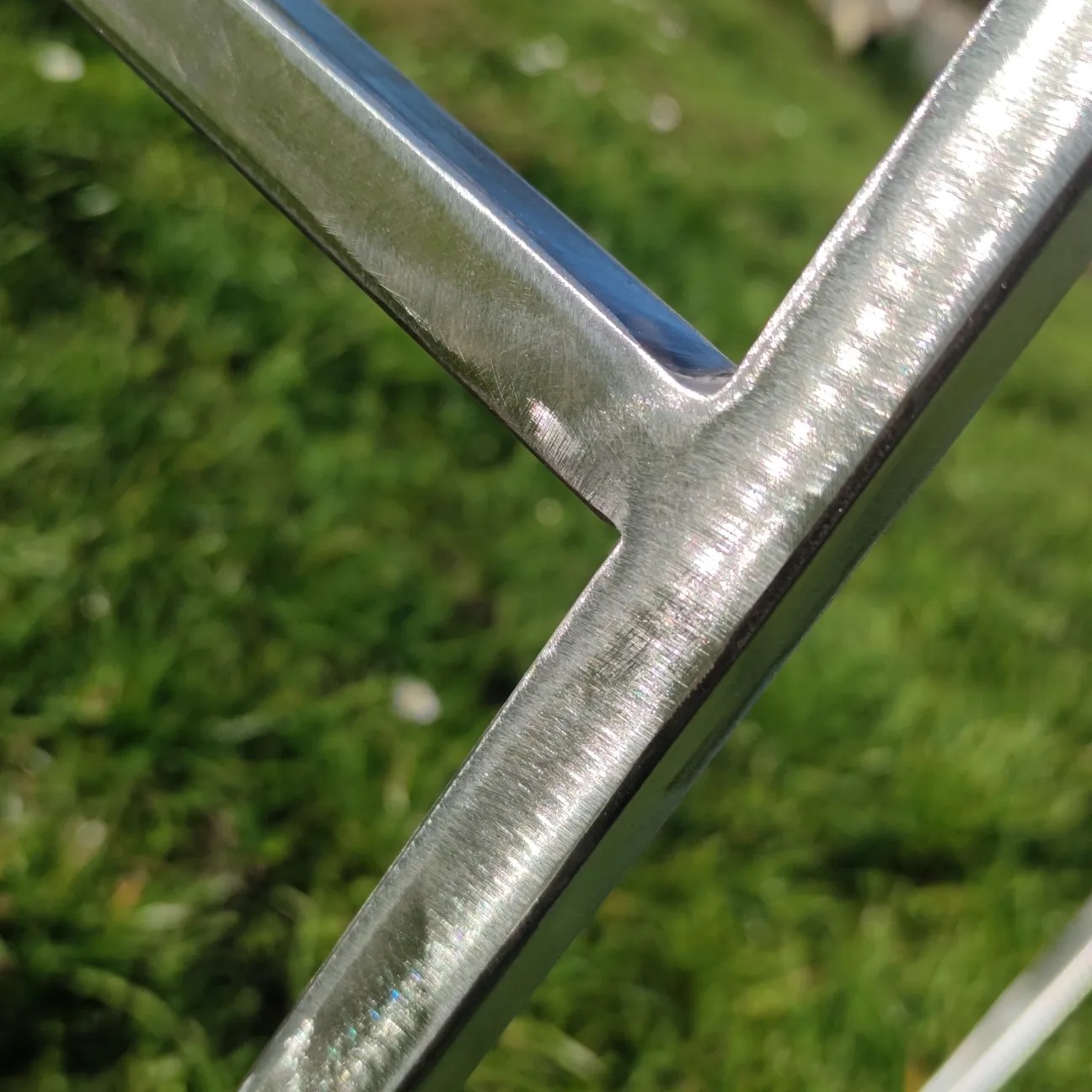 Spoina kątowa elementów metalowych o przekroju kwadratowym, widoczna faktura szlifowanego metalu na tle zielonej trawy.