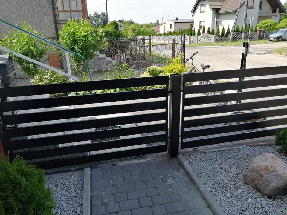 Nowoczesna, pozioma brama wjazdowa z czarnych, metalowych profili, widoczna od strony posesji, z brukowanym podjazdem i ozdobnym kamieniem na żwirze.