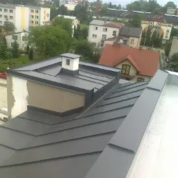 Szary dach z blachy na budynku z kominem wentylacyjnym, widok z góry na miasto w tle. Obróbki blacharskie wykończone w kolorze dachu.