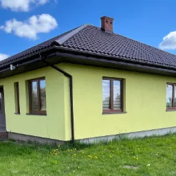 Żółty dom jednorodzinny z ciemnobrązowym dachem, widok z zewnątrz w słoneczny dzień, z widocznym fragmentem tarasu z kolumnami i niskim, ceglanym kominem.