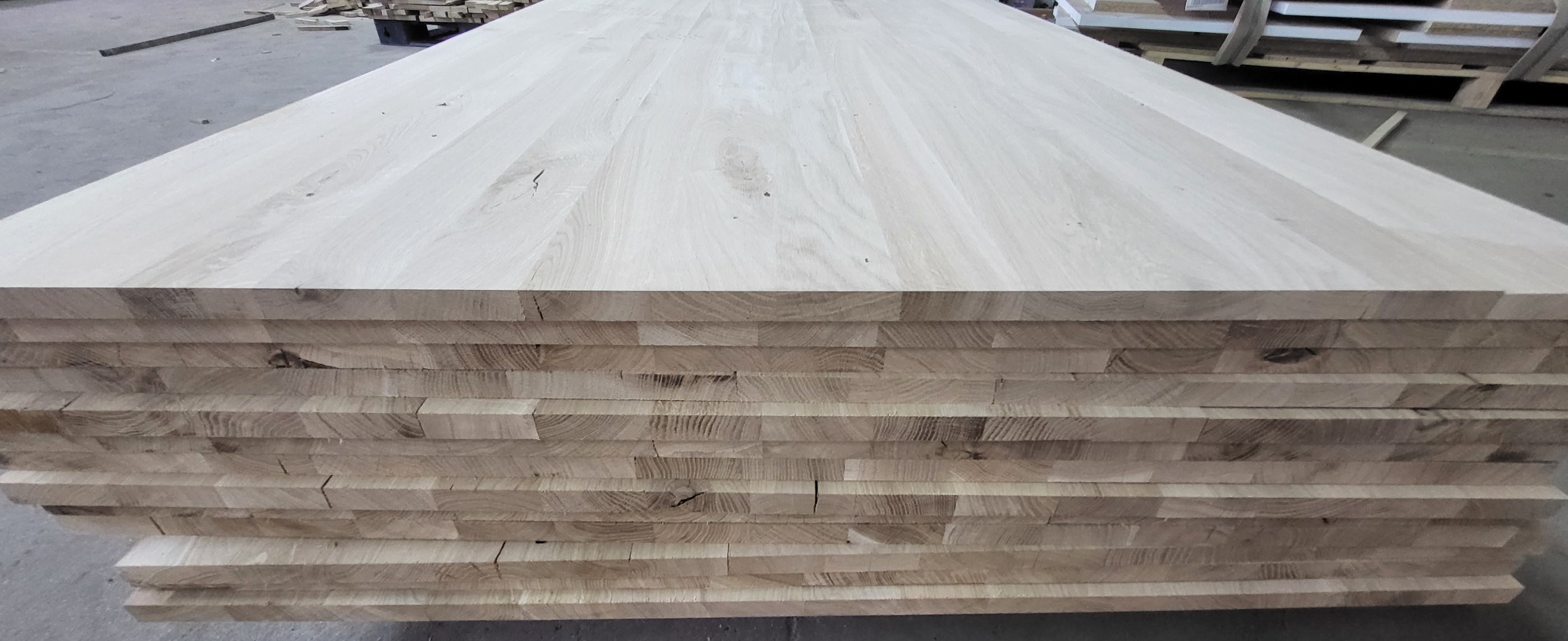 Stos jasnych, klejonych desek z widocznym usłojeniem, ułożonych warstwami w hali produkcyjnej.