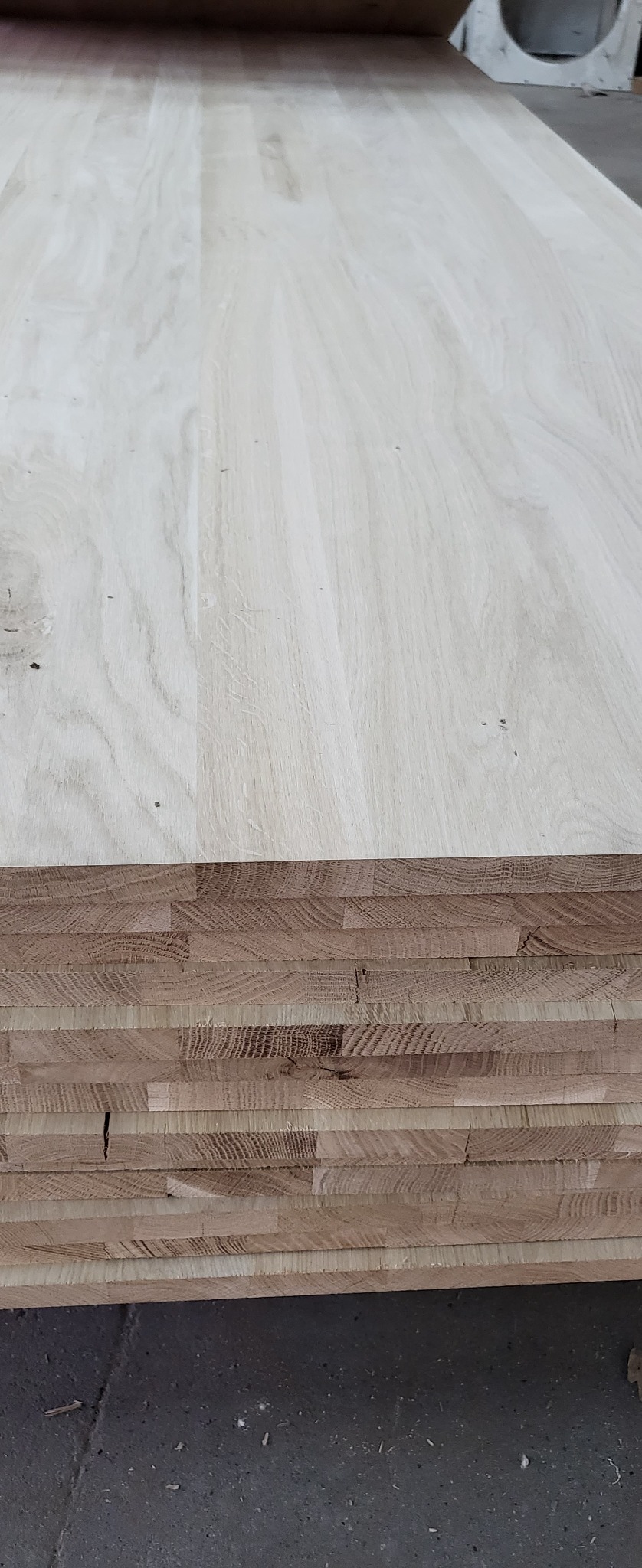 Stos jasnych, klejonych desek drewnianych z widocznym przekrojem poprzecznym, ukazującym strukturę klejenia na tle szarej, betonowej podłogi.