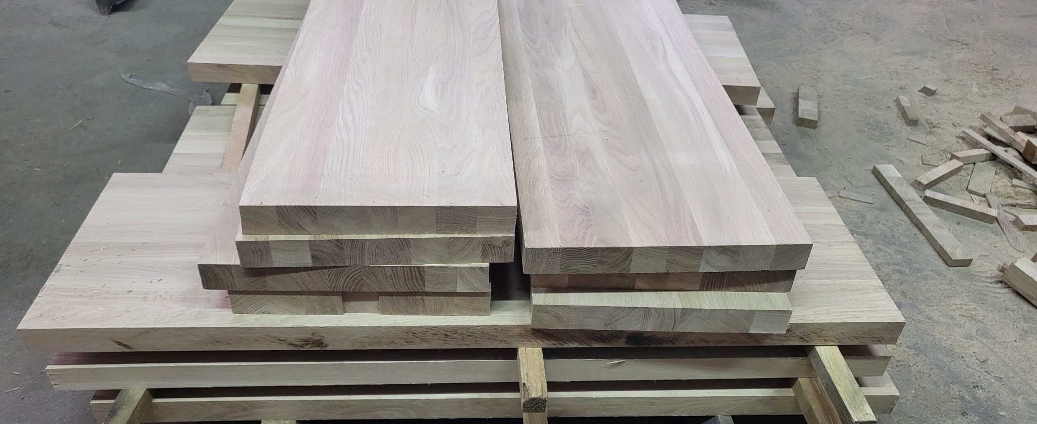 Deski klejone warstwowo, ułożone na palecie w warsztacie, z widocznymi wiórami i odpadami drewna w tle.