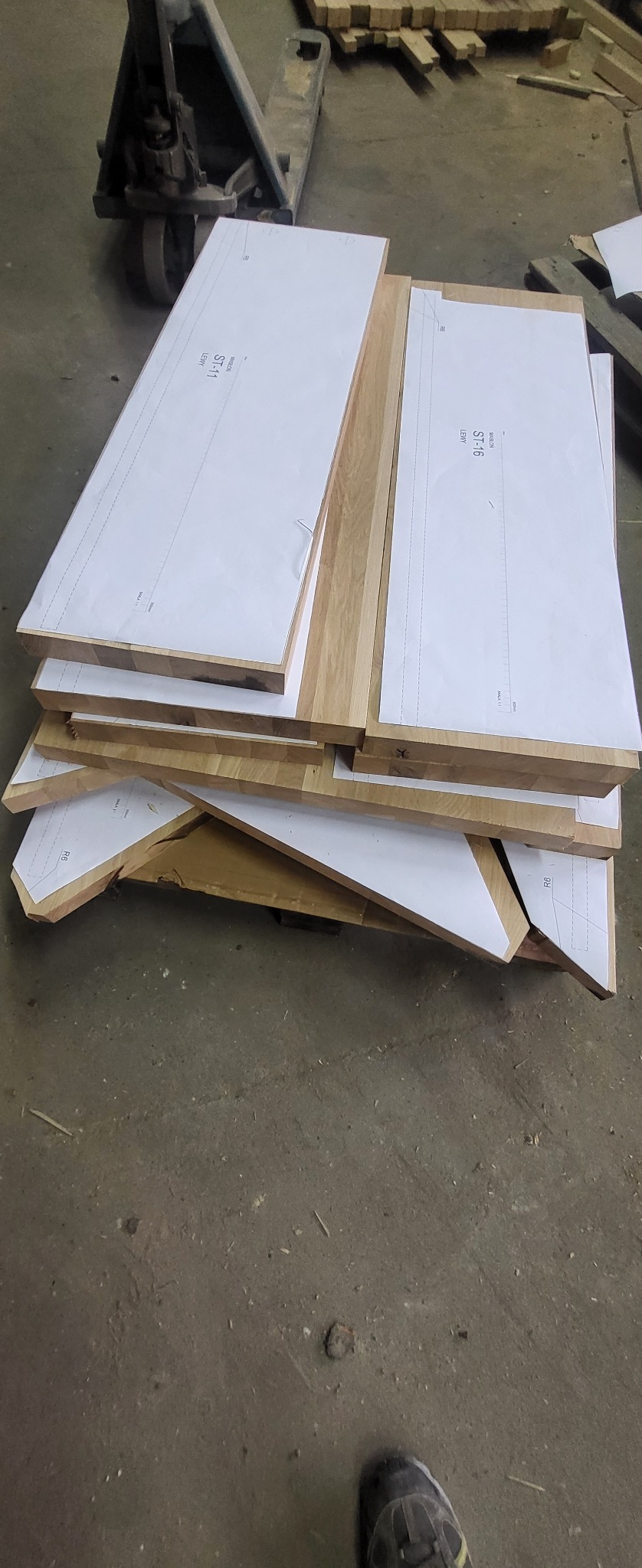 Stos drewnianych elementów z naklejonymi szablonami do wycinania, widoczny wózek paletowy w tle.