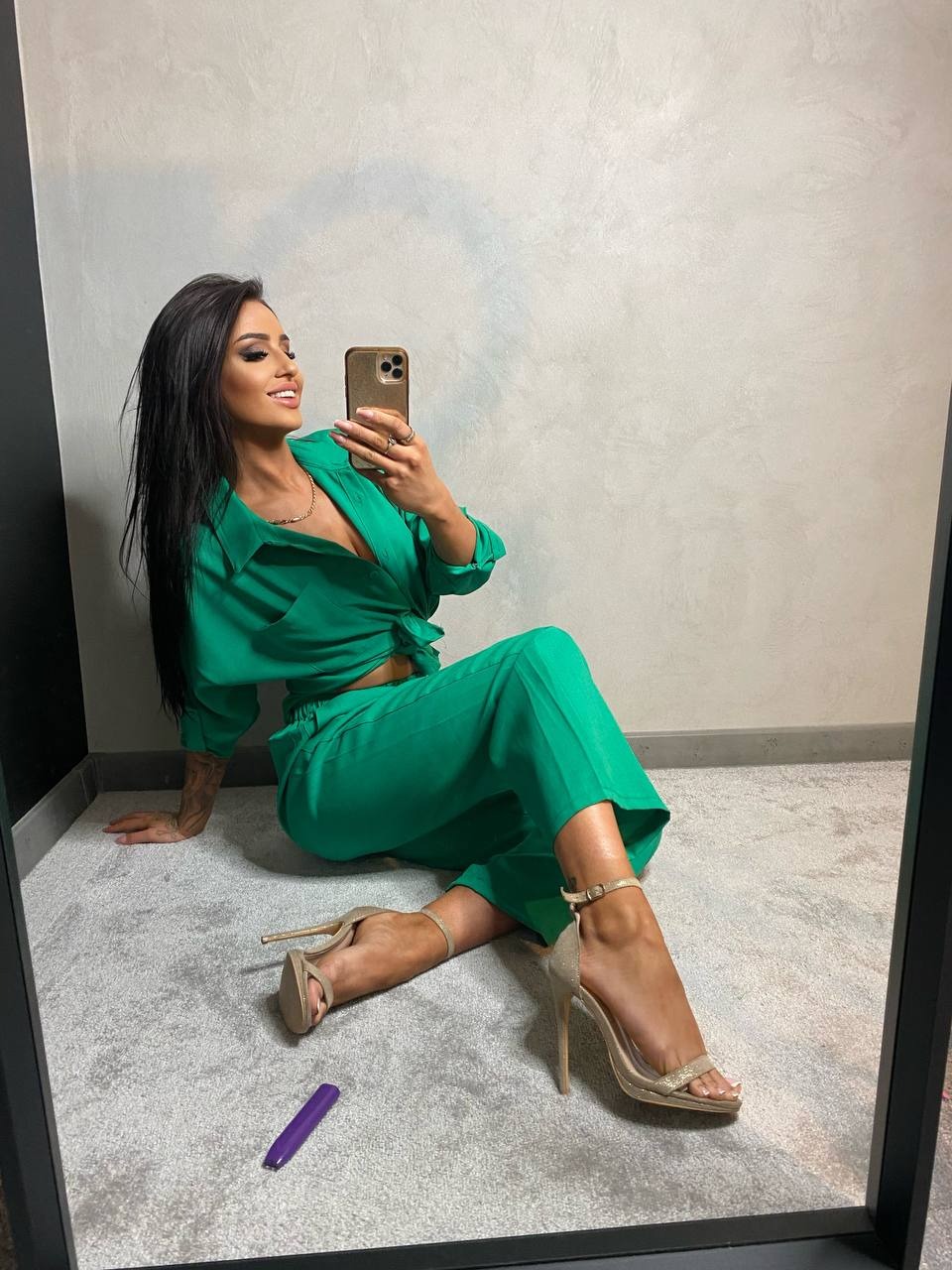 Kobieta z długimi, ciemnymi włosami pozuje do selfie w lustrze, ubrana w luźny, zielony komplet i złote sandały na obcasie, siedząc na szarym dywanie, obok fioletowej kapsułki.