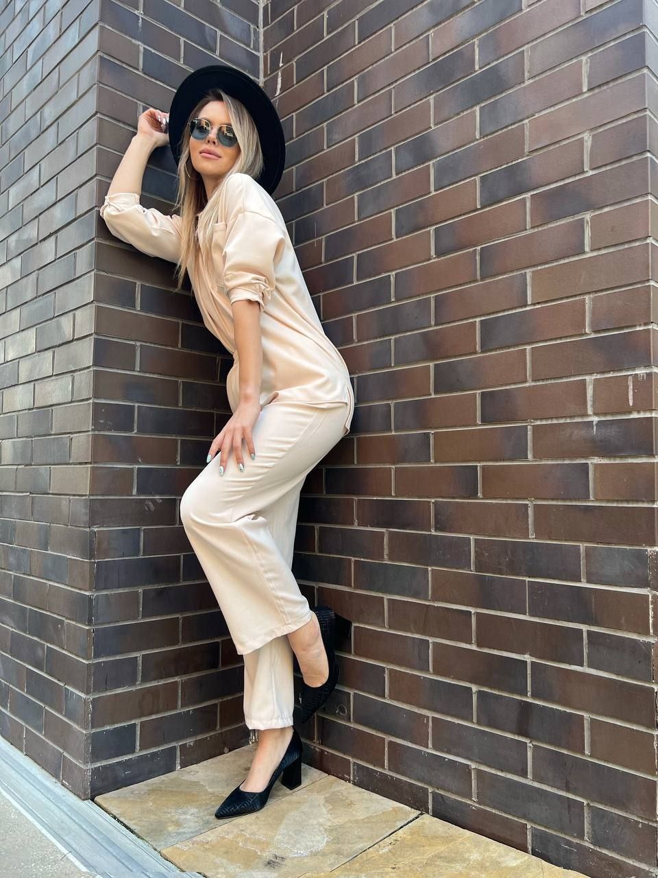Kobieta w beżowym kombinezonie, czarnym kapeluszu i okularach przeciwsłonecznych pozuje oparta o ceglaną ścianę. Widoczne czarne buty na obcasie z delikatnym wzorem.