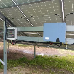 Panel słoneczny widziany od spodu, z widoczną konstrukcją wsporczą, skrzynką elektryczną i falownikiem z naklejką informacyjną, na tle trawy i ziemi.