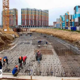 Prace zbrojarskie na budowie w Gdańsku: widok na siatkę zbrojeniową fundamentów, z pracownikami w kaskach i pomarańczowych koszulach, na tle wykopu i wielobarwnych budynków mieszkalnych.