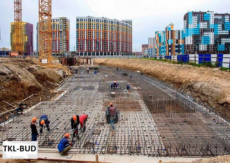 Prace zbrojarskie na budowie w Gdańsku: widok na siatkę zbrojeniową fundamentów, z pracownikami w kaskach i pomarańczowych koszulach, na tle wykopu i wielobarwnych budynków mieszkalnych.