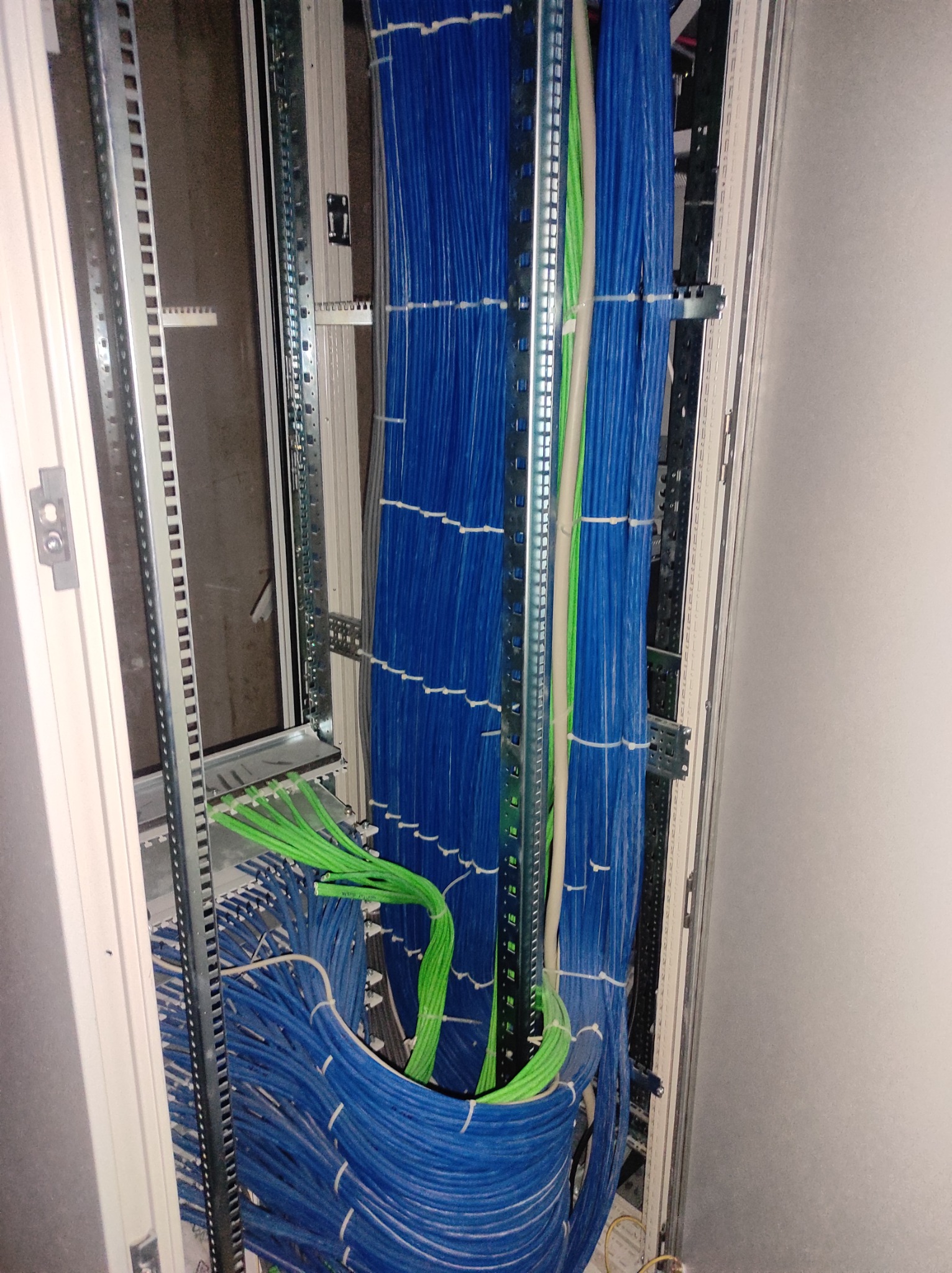 Szafa serwerowa z uporządkowanymi kablami sieciowymi w kolorze niebieskim i zielonym, zamocowanymi do metalowej ramy z widocznymi uchwytami i listwami montażowymi.