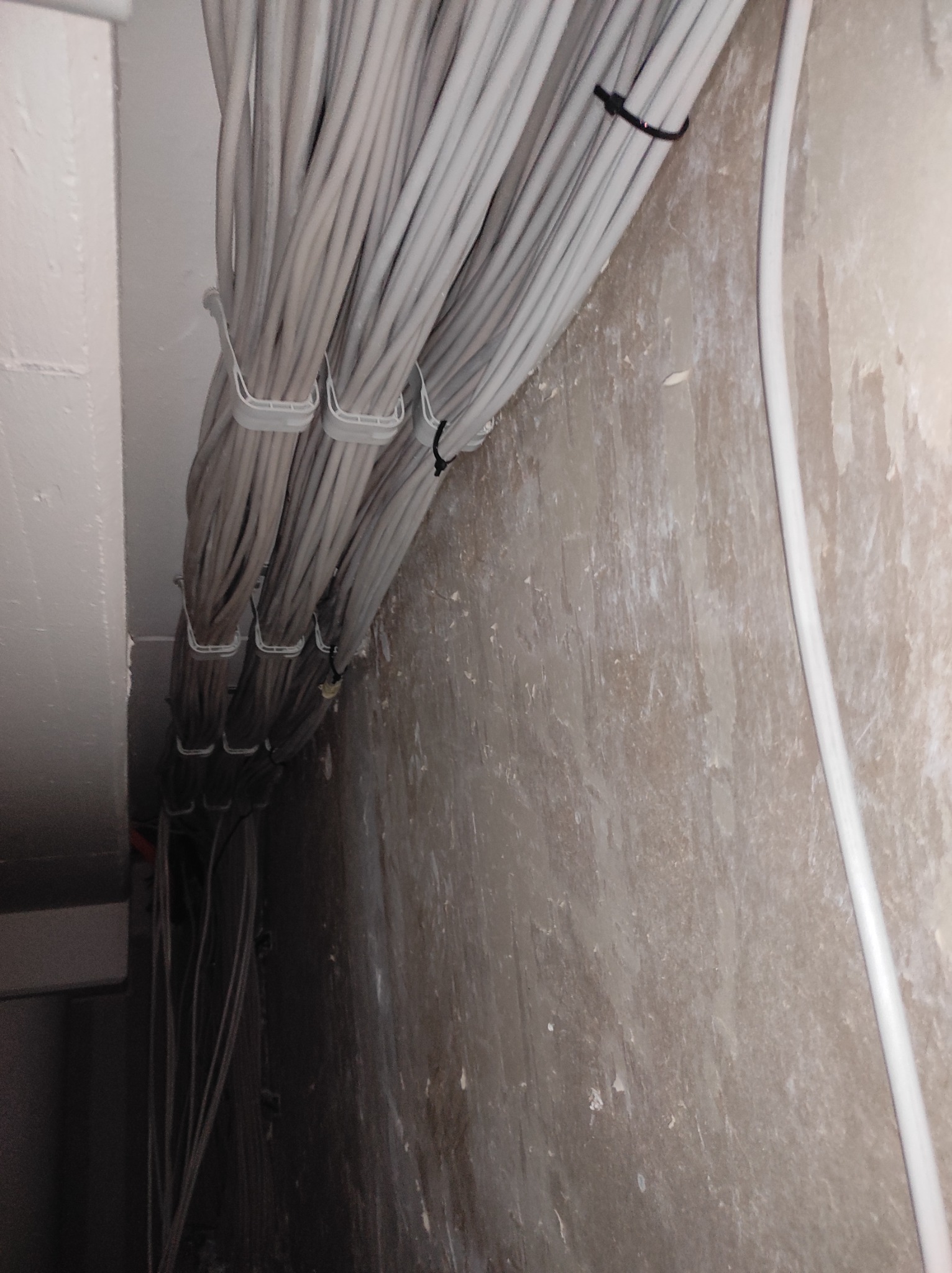 Pęki szarych kabli elektrycznych przymocowane do sufitu za pomocą białych i czarnych opasek zaciskowych na tle surowej ściany.