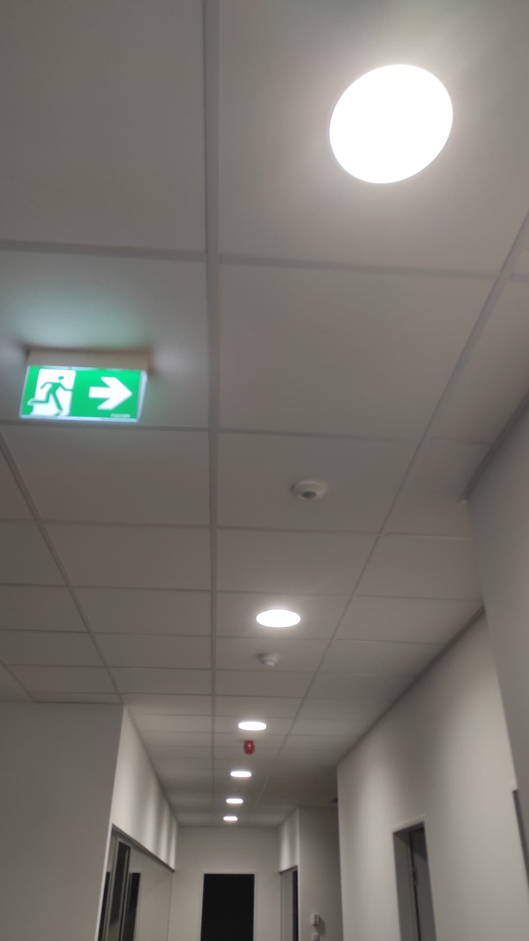 Korytarz biurowy z sufitem podwieszanym, oświetleniem LED i zielonym znakiem ewakuacyjnym z piktogramem osoby biegnącej.