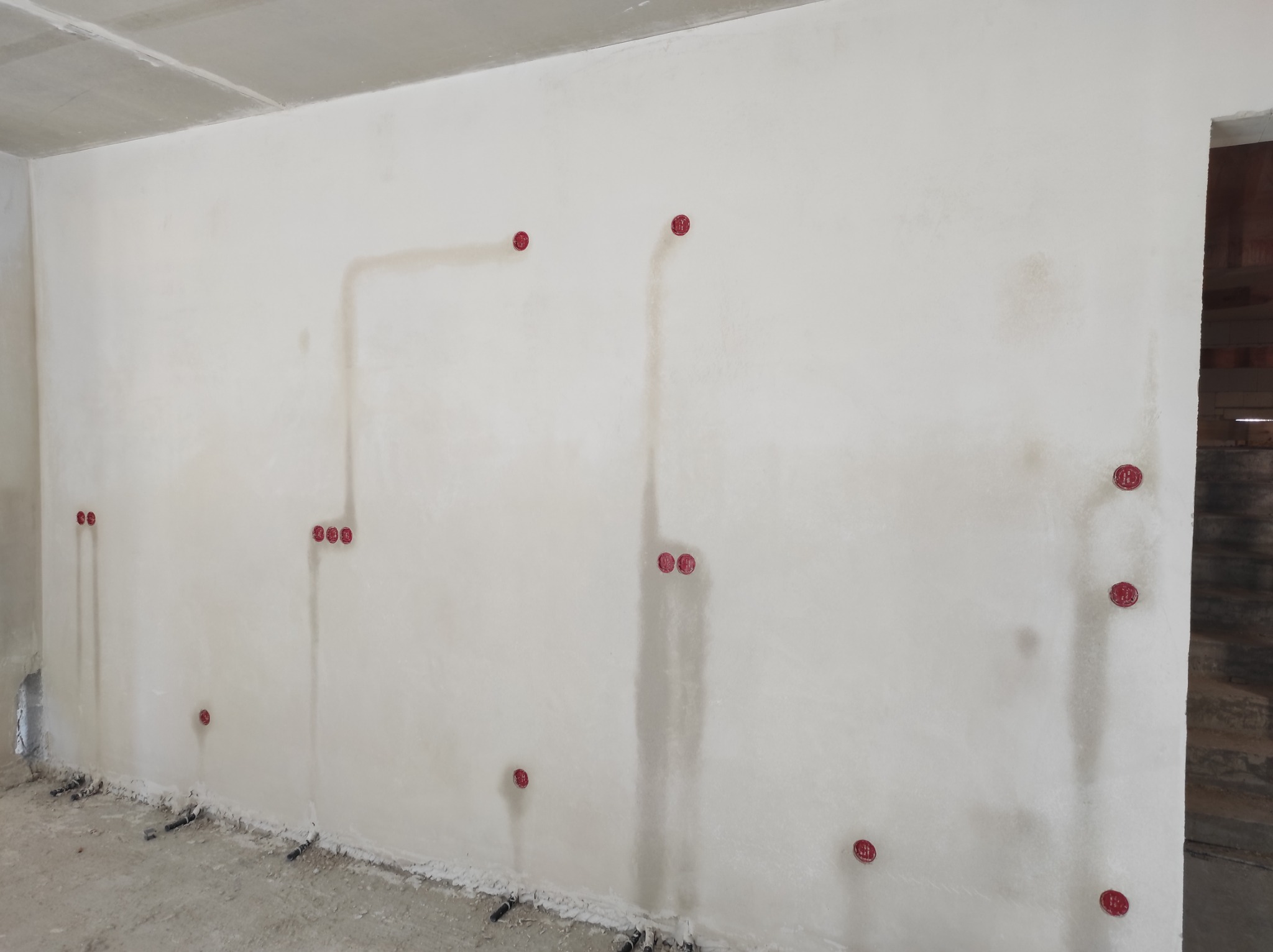 Świeżo otynkowana ściana z widocznymi, czerwonymi puszkami instalacyjnymi, przygotowana pod instalację elektryczną w nowym budynku.