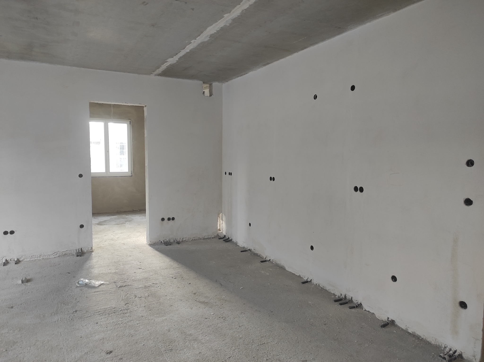 Surowy stan wnętrza z widocznymi puszkami elektrycznymi w ścianach i wystającymi rurkami przy podłodze, betonowy sufit, okno w tle.