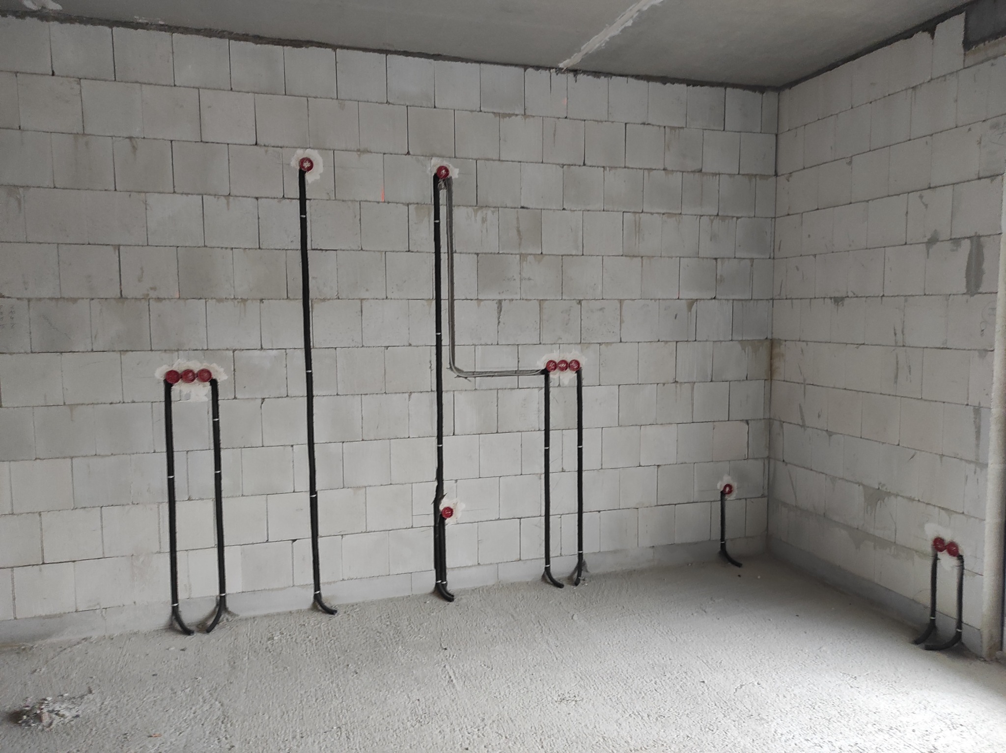 Wnętrze w stanie surowym z widocznymi przewodami elektrycznymi wyprowadzonymi ze ściany z betonu komórkowego, zabezpieczonymi czerwonymi puszkami, przygotowane do dalszych prac wykończeniowych.