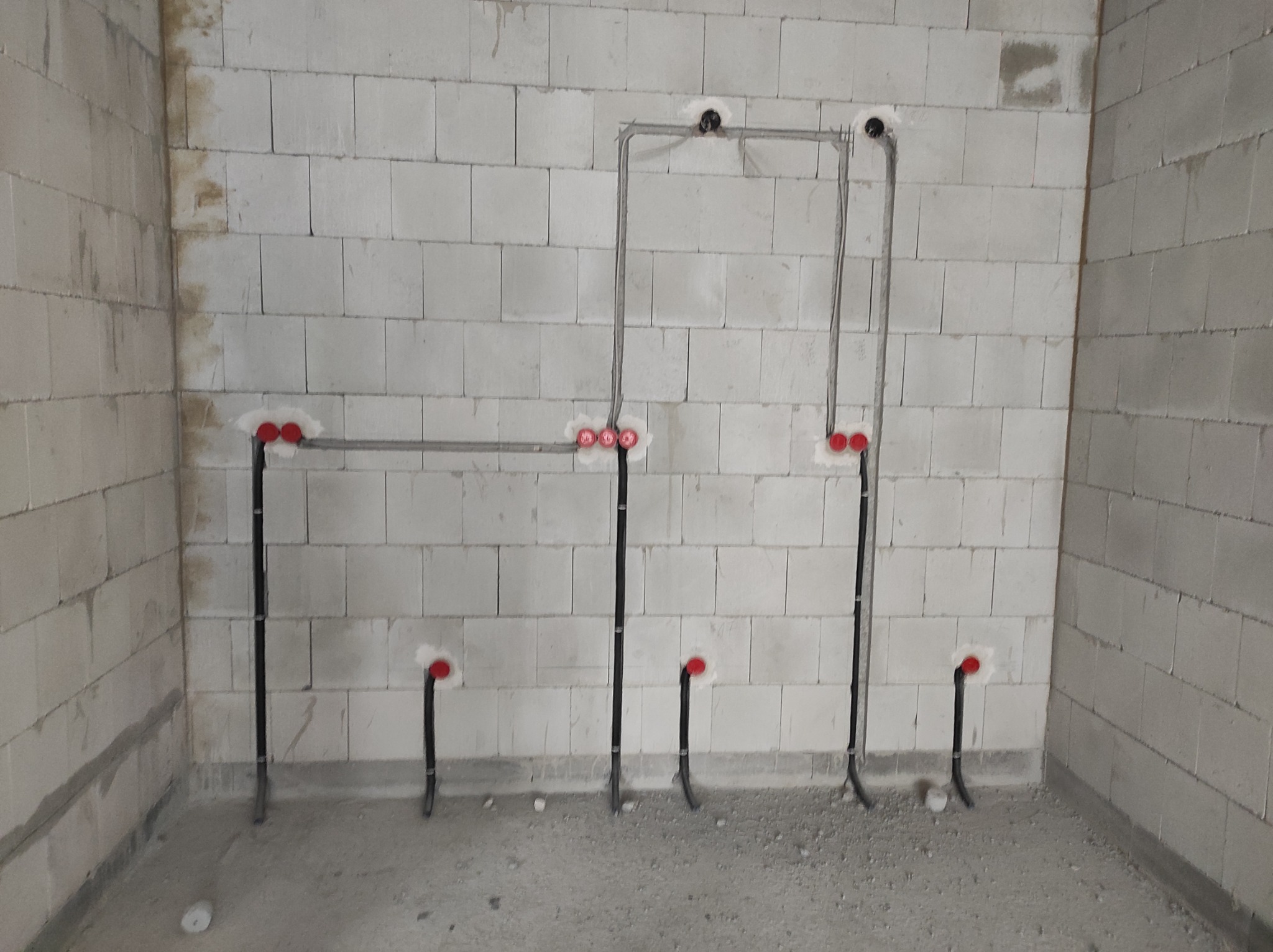 Surowe ściany z betonu komórkowego z widoczną, częściowo zamontowaną instalacją elektryczną: pionowe i poziome rury oraz puszki z czerwonymi zaślepkami.