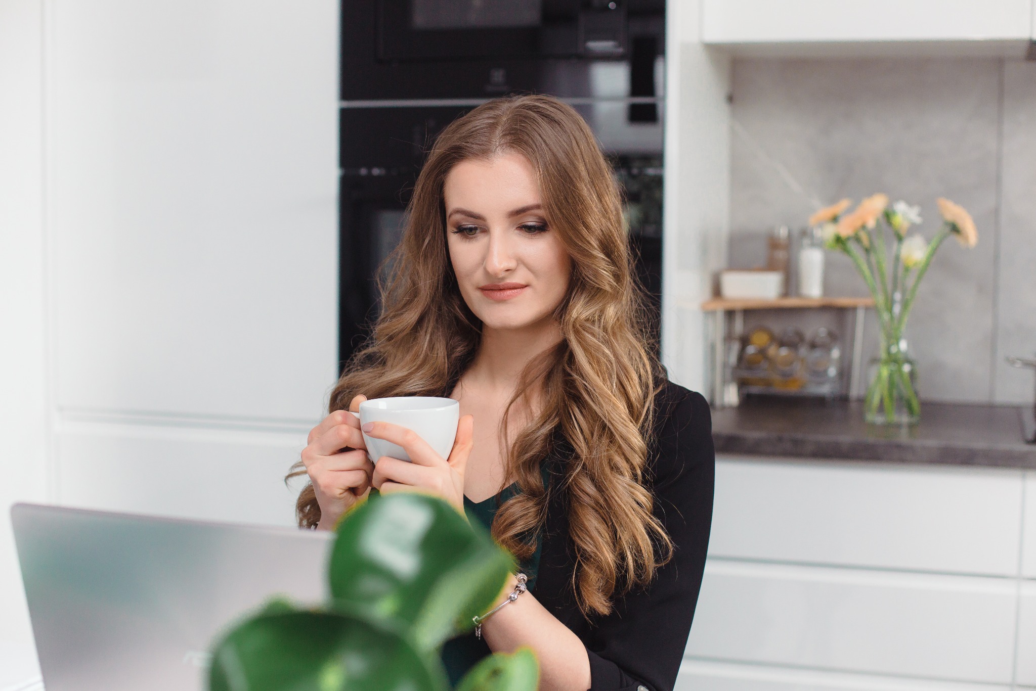 Kobieta z długimi włosami w Częstochowie, pijąca kawę z białego kubka, siedząc przed laptopem w jasnej kuchni z kwiatami w tle, podczas konsultacji online.
