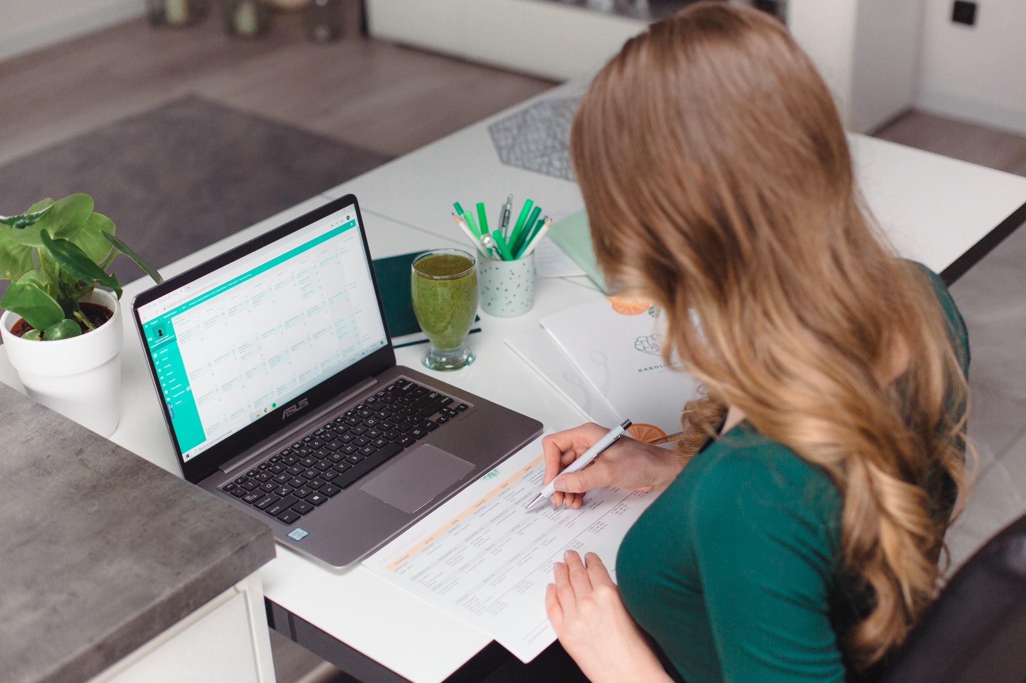 Kobieta analizuje plan żywieniowy na papierze, obok laptop z otwartym programem, szklanka zielonego smoothie i roślina doniczkowa na biurku.