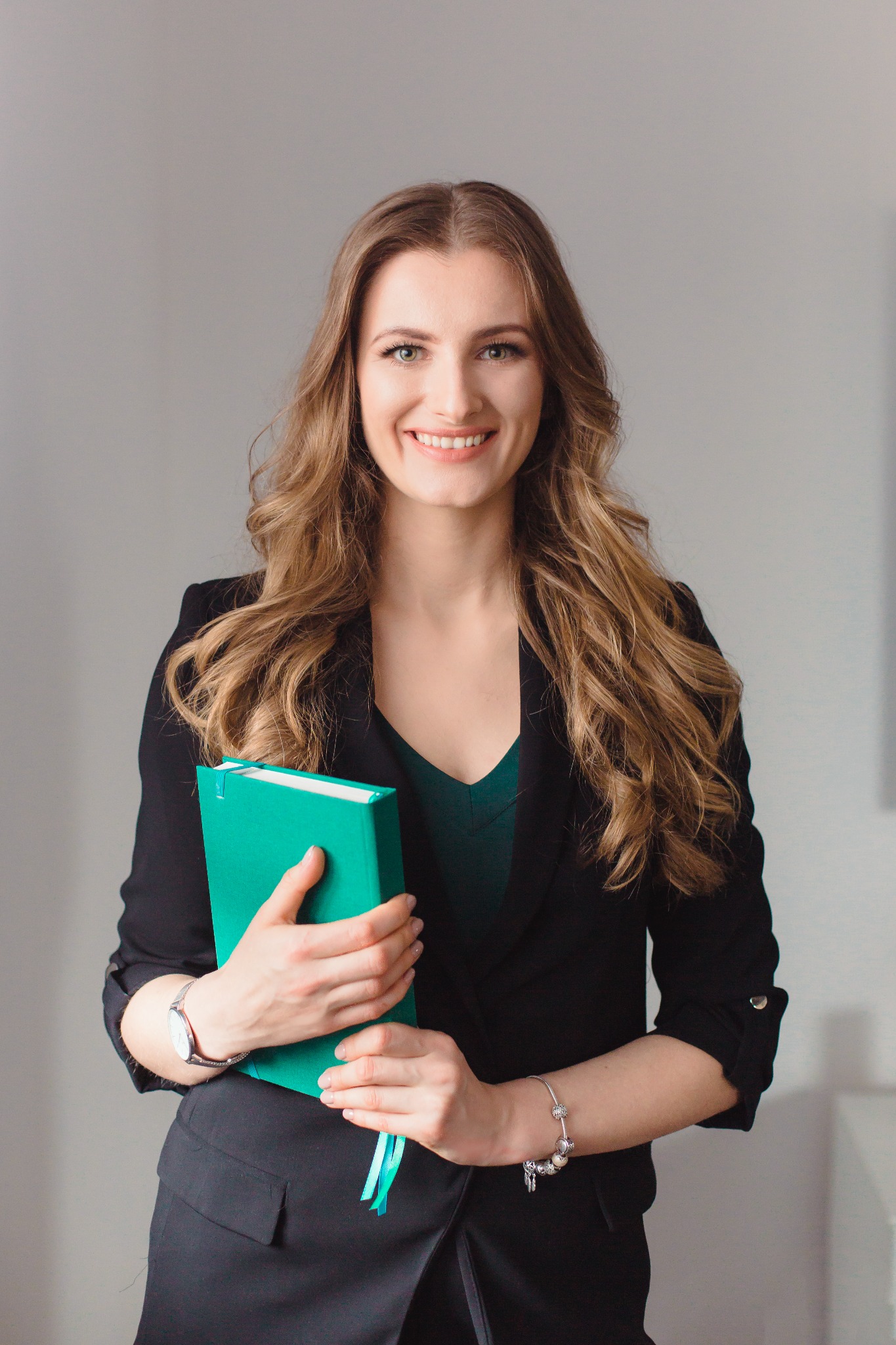 Uśmiechnięta kobieta w czarnej marynarce trzyma zielony notatnik z zakładkami, widoczny zegarek na ręku i bransoletka z charmsami.