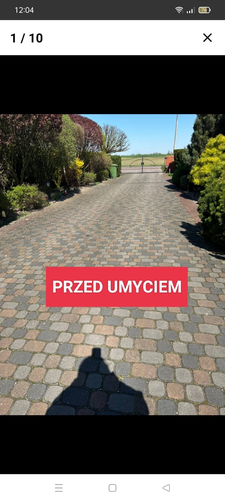 Długa, dwukolorowa (szaro-brązowa) kostka brukowa na podjeździe do posesji, widok w kierunku bramy wjazdowej i zieleni, z cieniem fotografa w dolnej części kadru, z czerwonym napisem 'PRZED...