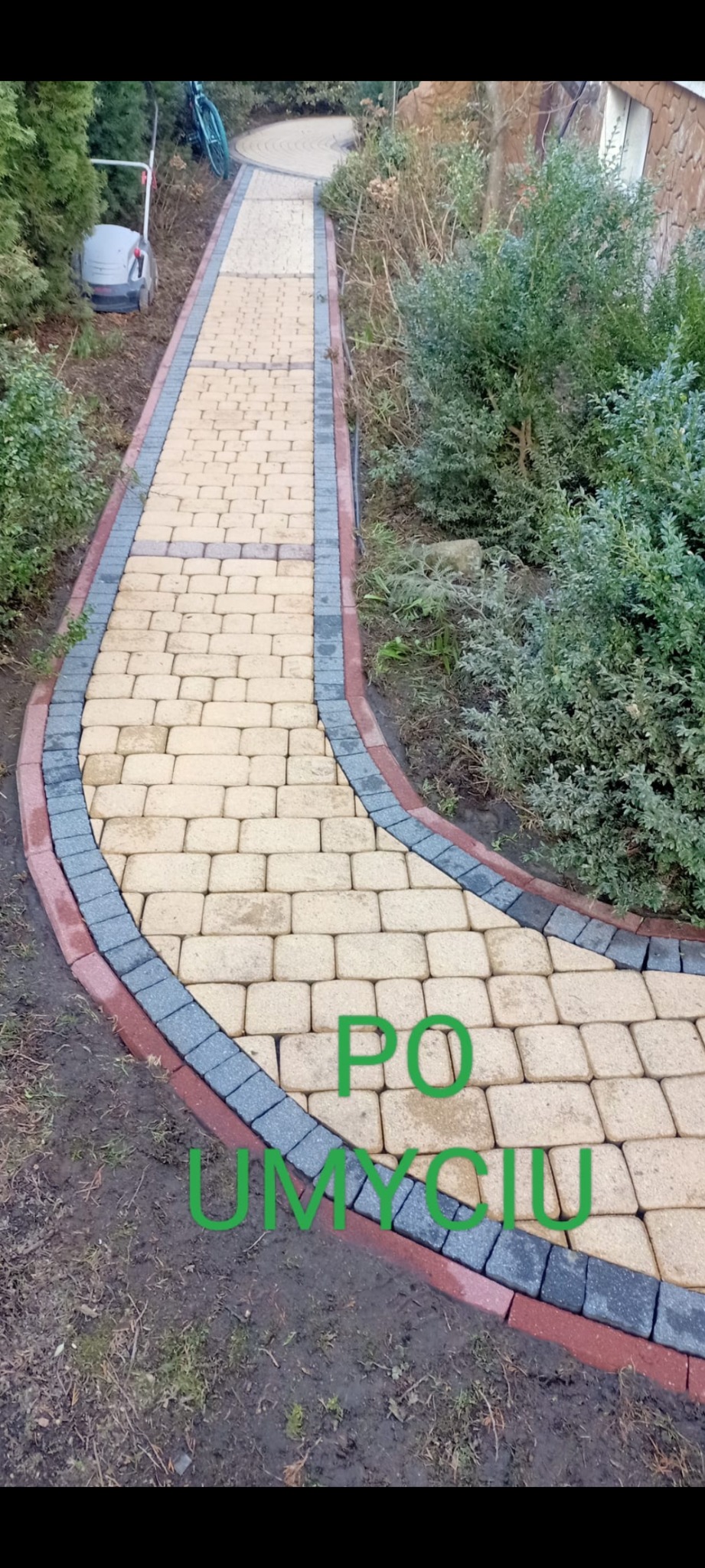 Ścieżka z kostki brukowej po umyciu, z obrzeżem z kostki w kolorach szarym i czerwonym, widoczna trawa i krzewy po bokach, w tle kosiarka i rower.