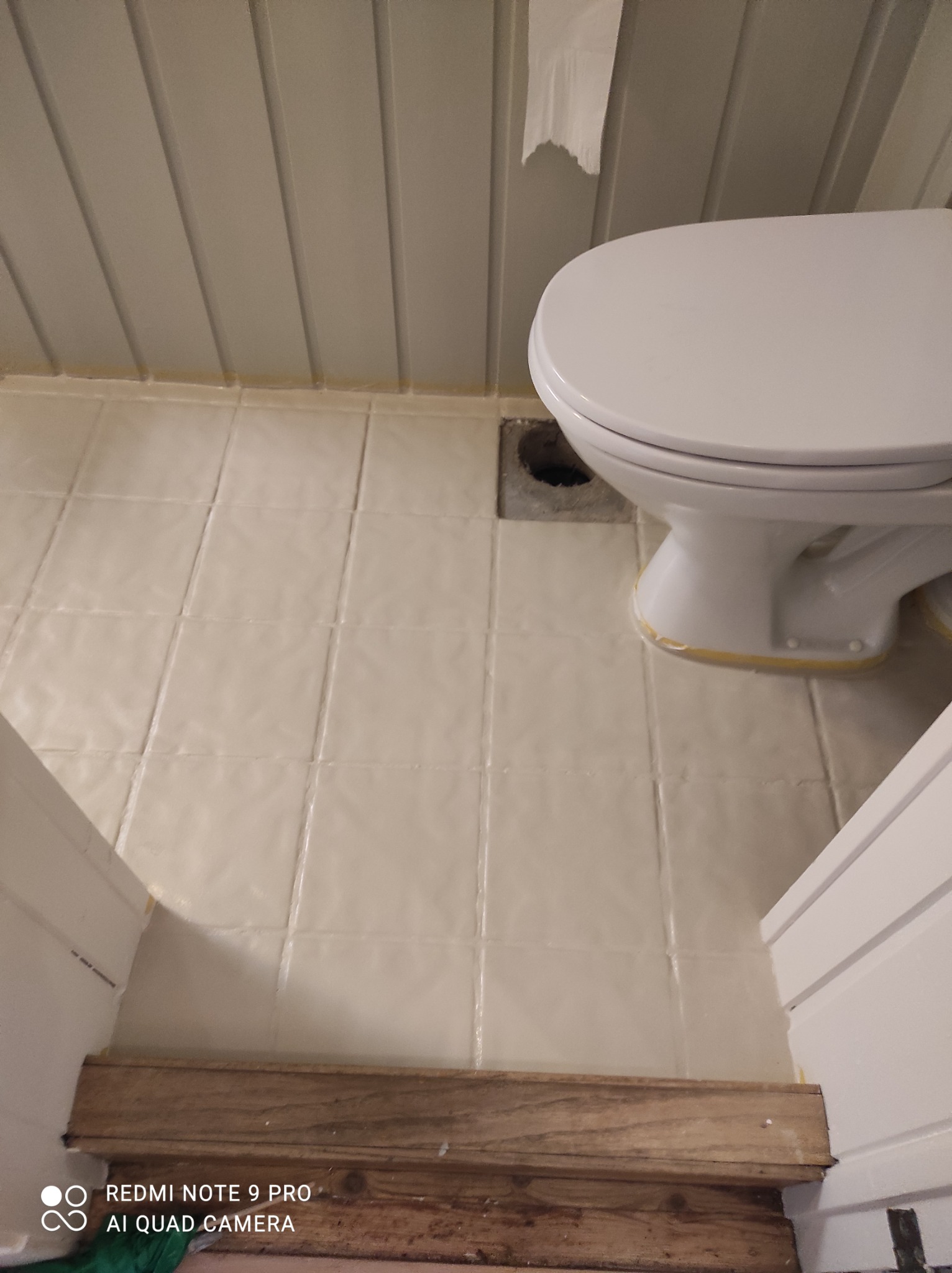 Łazienka po malowaniu, widoczne świeżo pomalowane płytki podłogowe i ściany z boazerią, zabezpieczona taśmą toaleta, drewniany próg.