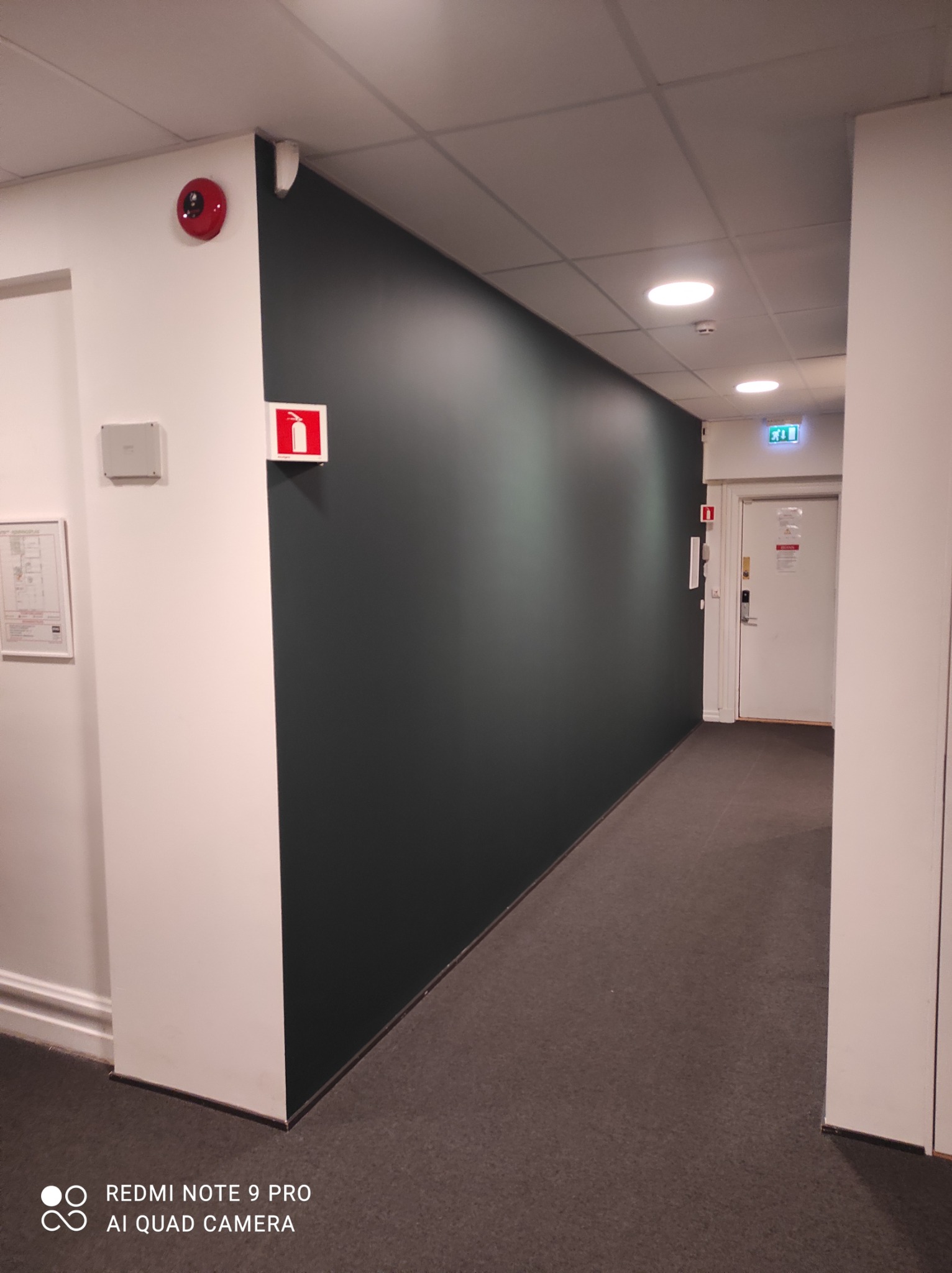 Korytarz biurowy z ciemnozieloną ścianą, białymi elementami architektonicznymi, szarą wykładziną podłogową i znakami bezpieczeństwa.