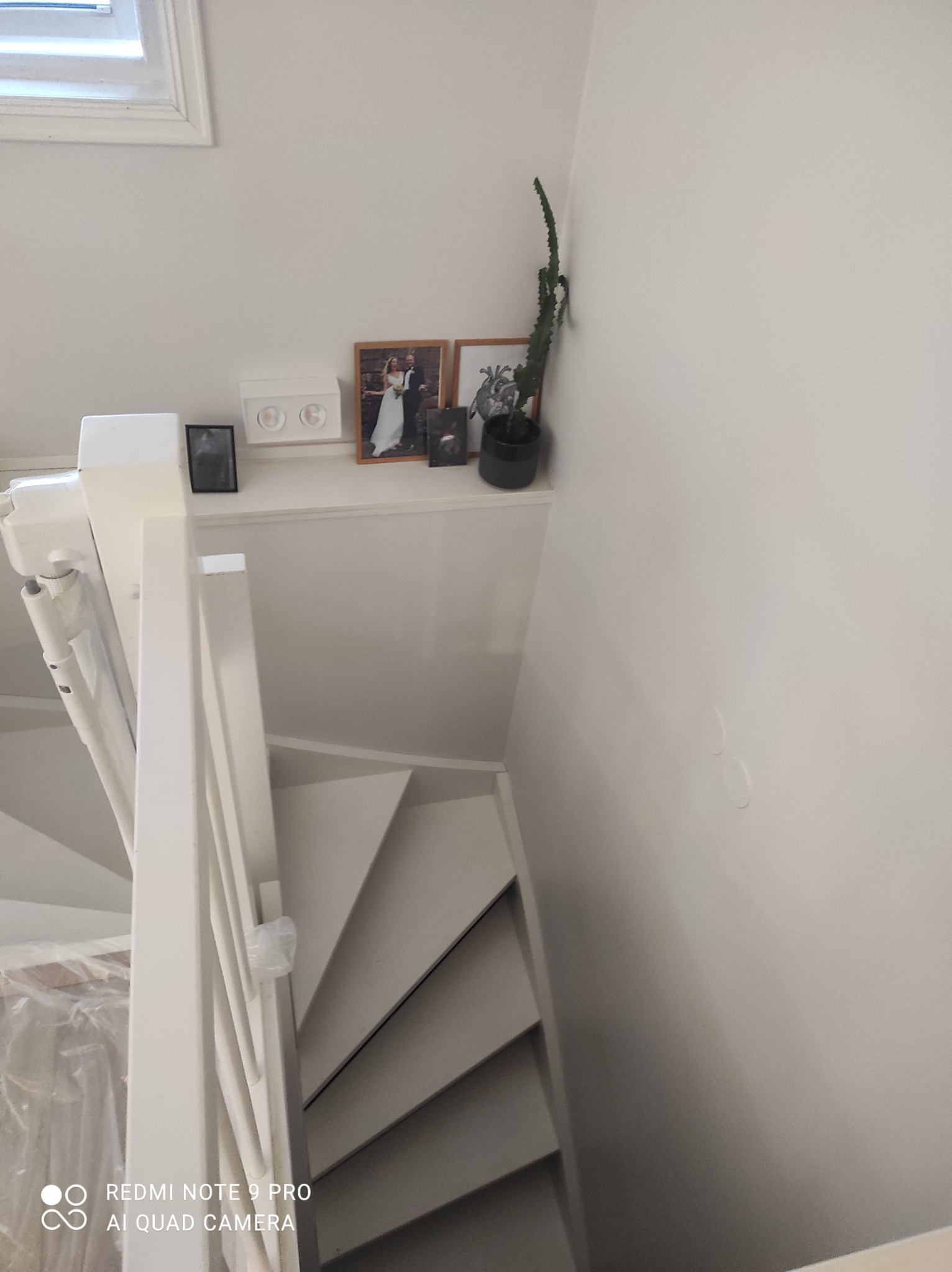 Białe schody pomalowane na biało, widok z góry, na ścianie półka z dekoracjami, w tym zdjęcia w ramkach i roślina doniczkowa, ściana świeżo pomalowana