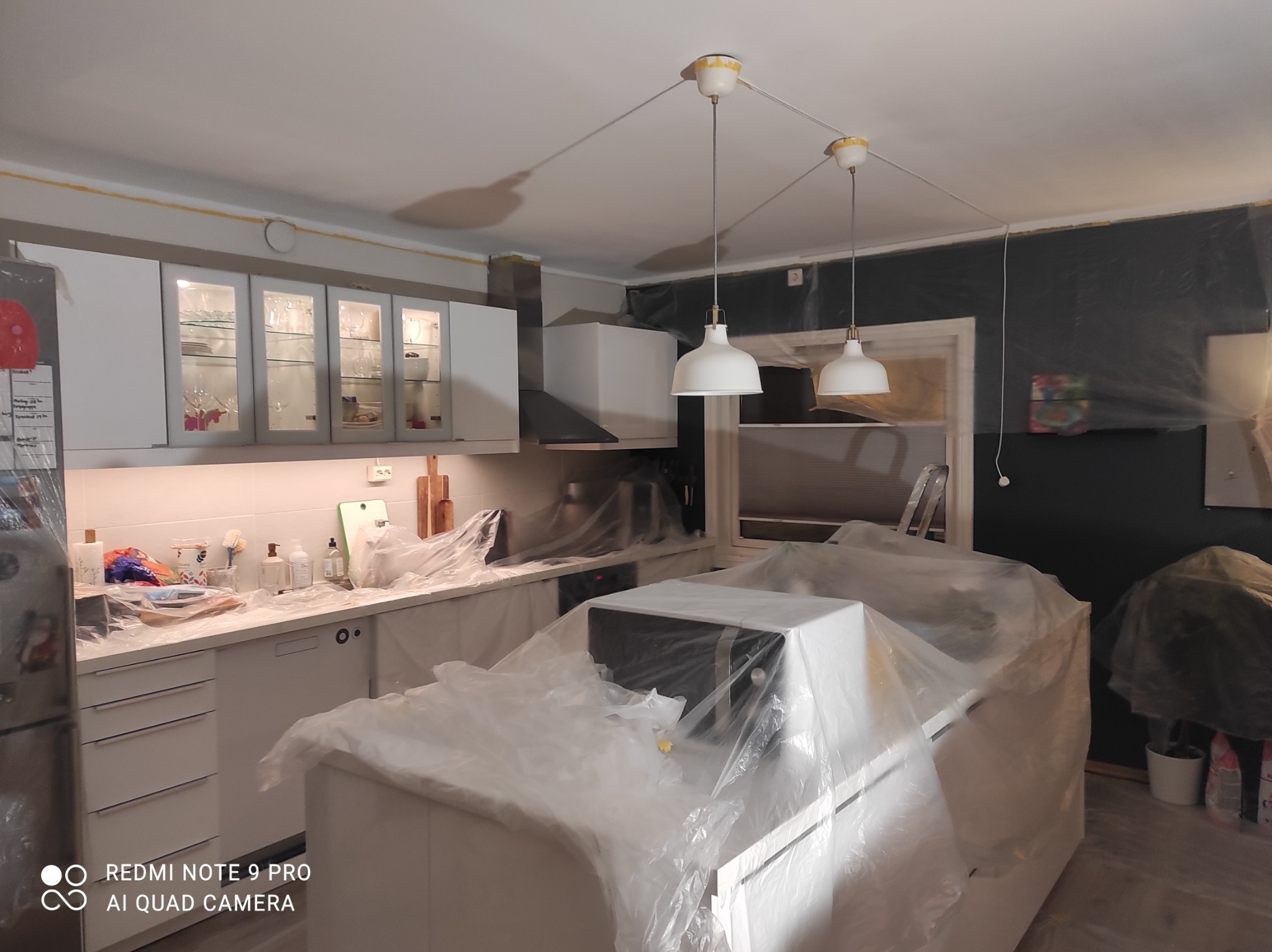 Kuchnia w trakcie remontu: meble i sprzęty zabezpieczone folią malarską, widoczne przygotowania do malowania ścian na ciemny kolor, trzy białe lampy wiszące.