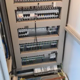 Otwarta szafa rozdzielcza z widocznymi zabezpieczeniami, przewodami i listwami zaciskowymi, z oznaczeniami na poszczególnych elementach, w szarej obudowie.