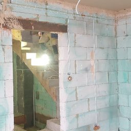 MIK-MONTAŻ Sp.z.o.o. - Wnętrze w trakcie remontu z widocznymi przewodami elektrycznymi w korytkach na ścianie z bloczków, z otworami na puszki elektryczne i instalacją schodów w tle.