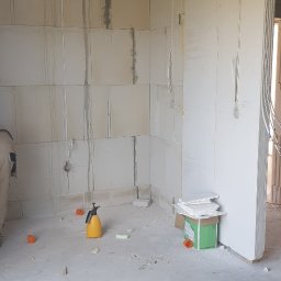 MIK-MONTAŻ Sp.z.o.o. - Wnętrze surowego budynku w trakcie instalacji elektrycznej; widoczne przewody elektryczne prowadzone pod sufitem i wzdłuż ścian z betonu komórkowego, żółty opryskiwacz ciśnieniowy i kartonowe...