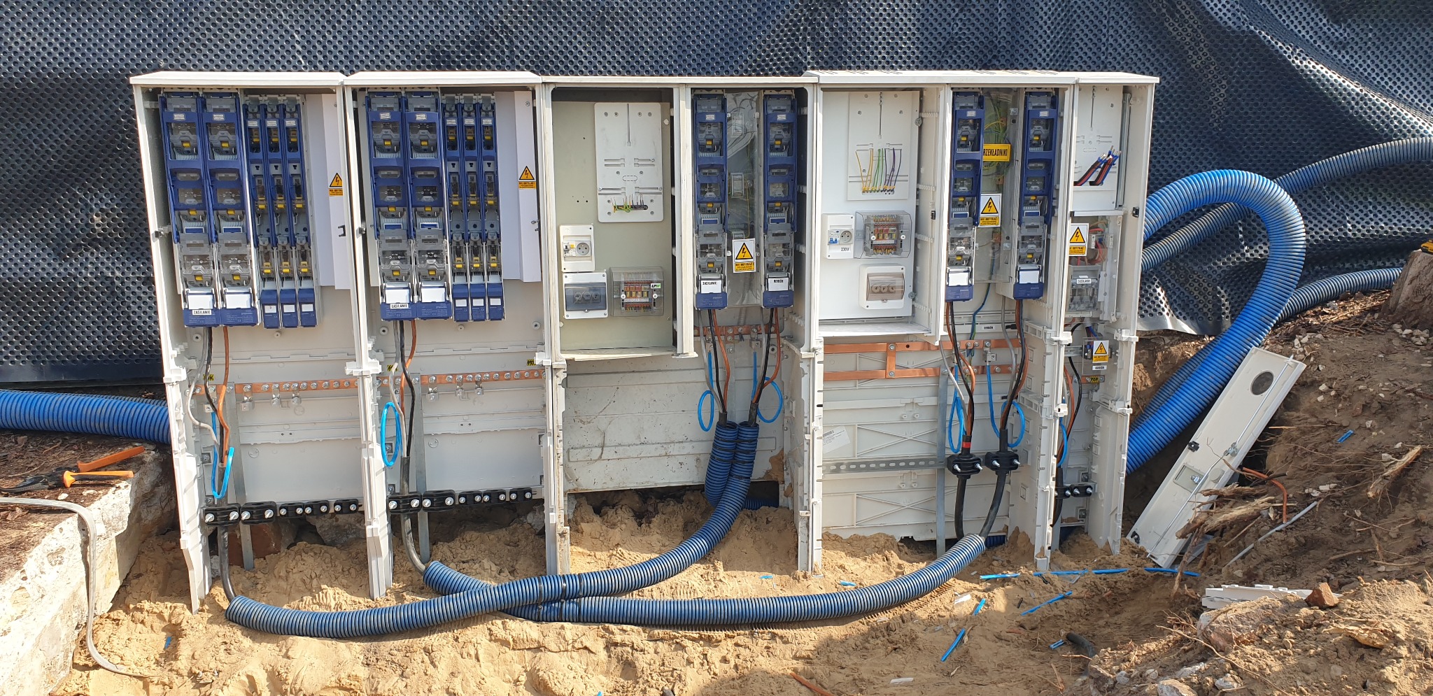 Szafki elektryczne przyłączeniowe w trakcie montażu w ziemi, widoczne kable i rury osłonowe, tabliczki ostrzegawcze, ujęcie z poziomu gruntu.