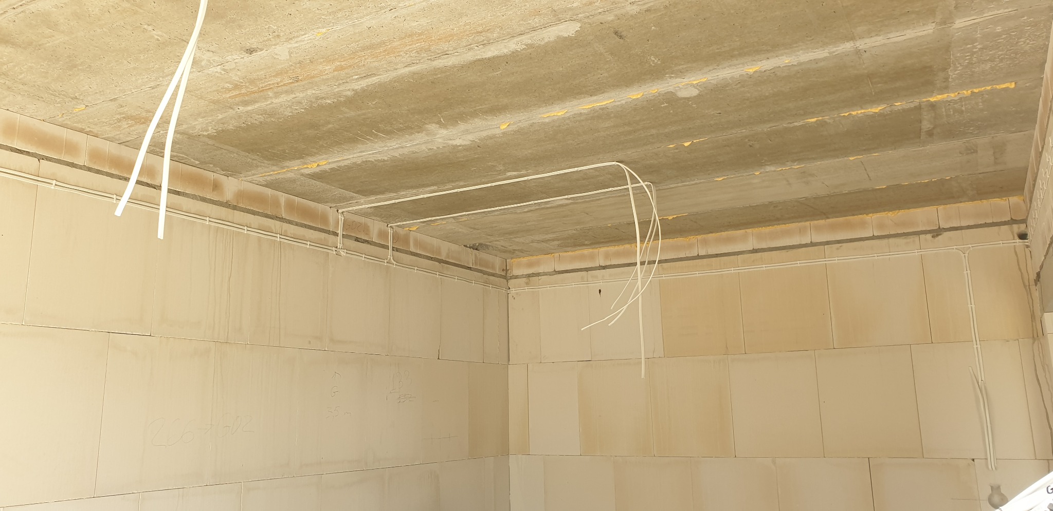 Wewnętrzna instalacja elektryczna w surowym pomieszczeniu z betonowym stropem i ścianami z bloczków, widoczne przewody elektryczne prowadzone po ścianach i suficie.