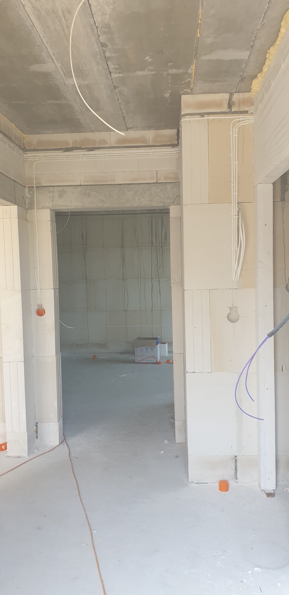 Surowy stan wnętrza w trakcie prac budowlanych z widocznymi przewodami w peszlach na ścianach i suficie, oraz wystającymi kablami ze ścian. W tle widoczny karton z materiałami budowlanymi.