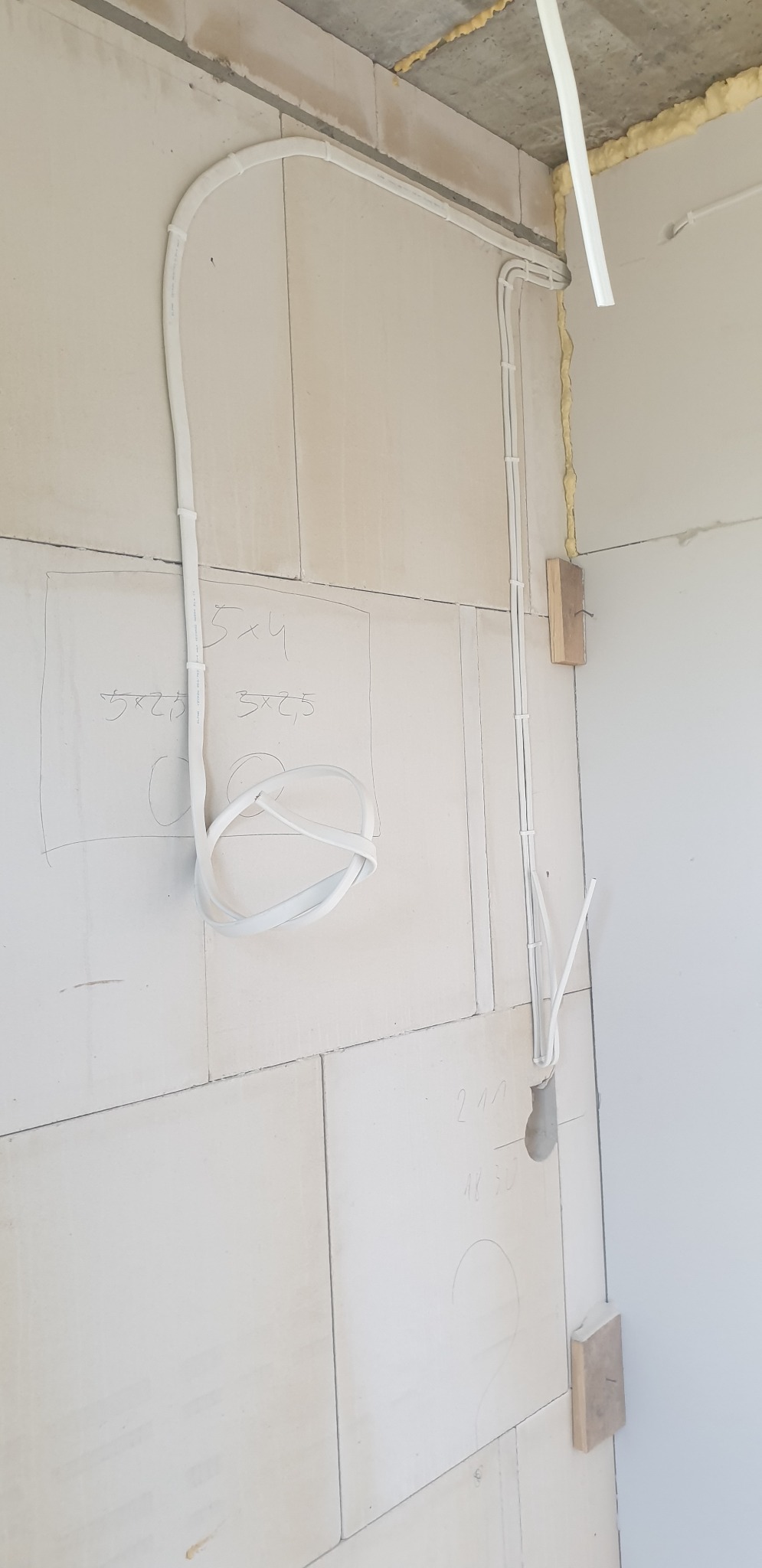 Białe przewody ułożone na ścianie z betonu komórkowego, tymczasowo przymocowane drewnianymi klockami, z widocznymi odręcznymi notatkami z wymiarami.