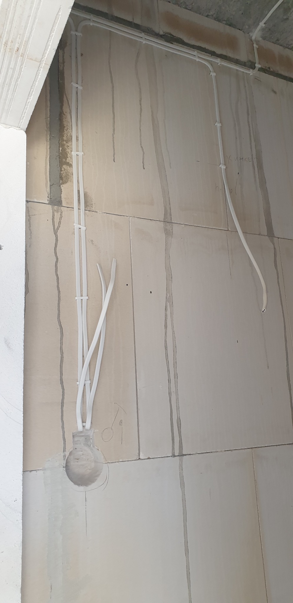 Wewnętrzne okablowanie elektryczne w białych rurkach PCV, przymocowane do surowej ściany z widocznymi zaciekami i oznaczeniami ołówkiem, prowadzące do otworu w ścianie.