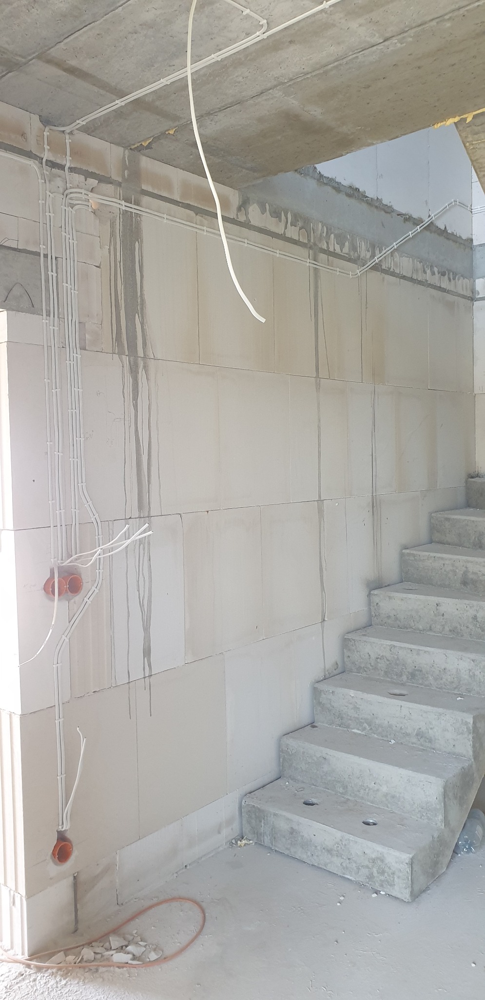 Surowe wnętrze w trakcie budowy z widocznymi przewodami w peszlach zamontowanymi na ścianie z bloczków betonowych oraz betonowymi schodami.