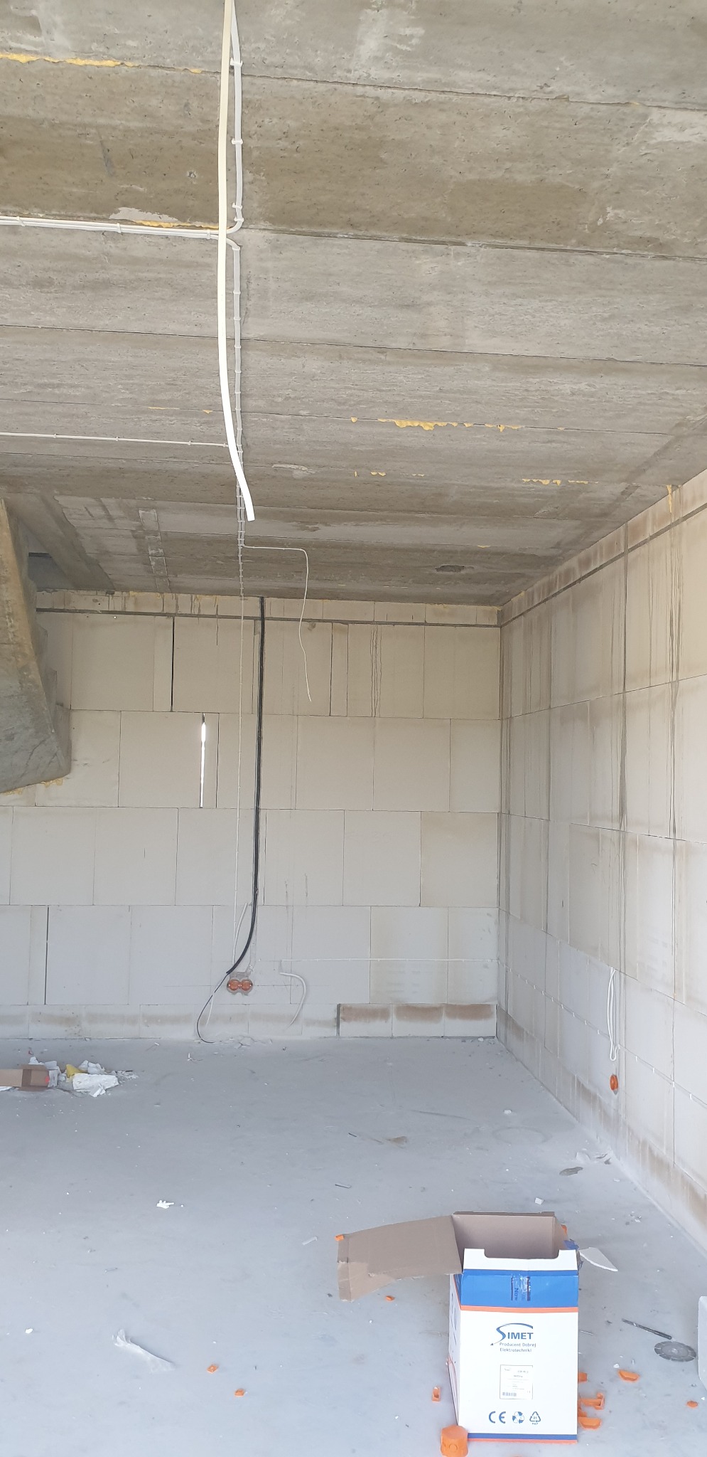 Surowy stan wnętrza: widoczne przewody elektryczne, puszki podtynkowe, ściany z betonu komórkowego i sufit z betonu. Na podłodze kartonowe pudełko i resztki materiałów budowlanych.