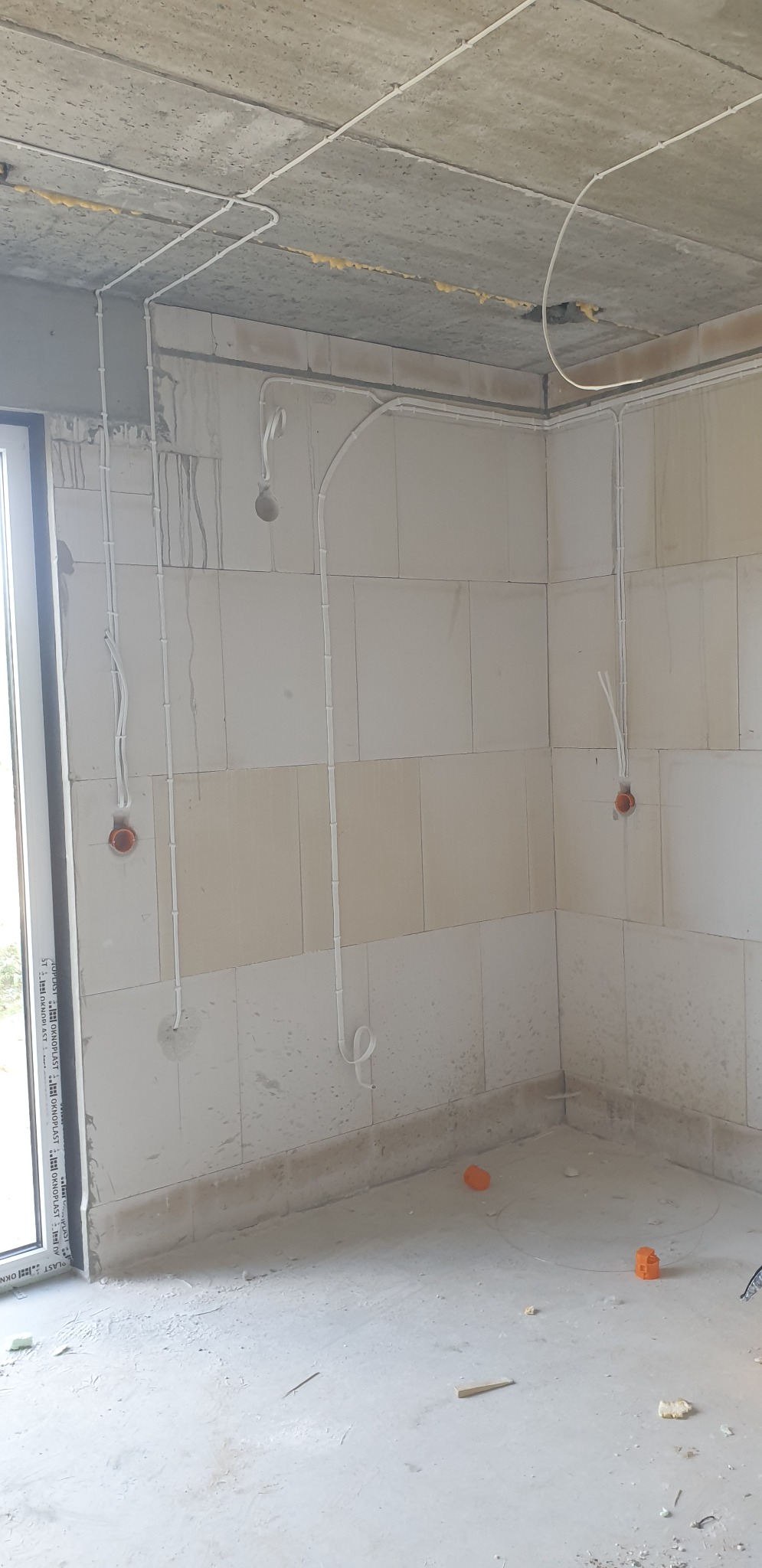 Wnętrze w stanie surowym z widoczną instalacją elektryczną: białe kable prowadzone po ścianach z bloczków i suficie, puszki pod gniazdka i wypust kabla na żarówkę.