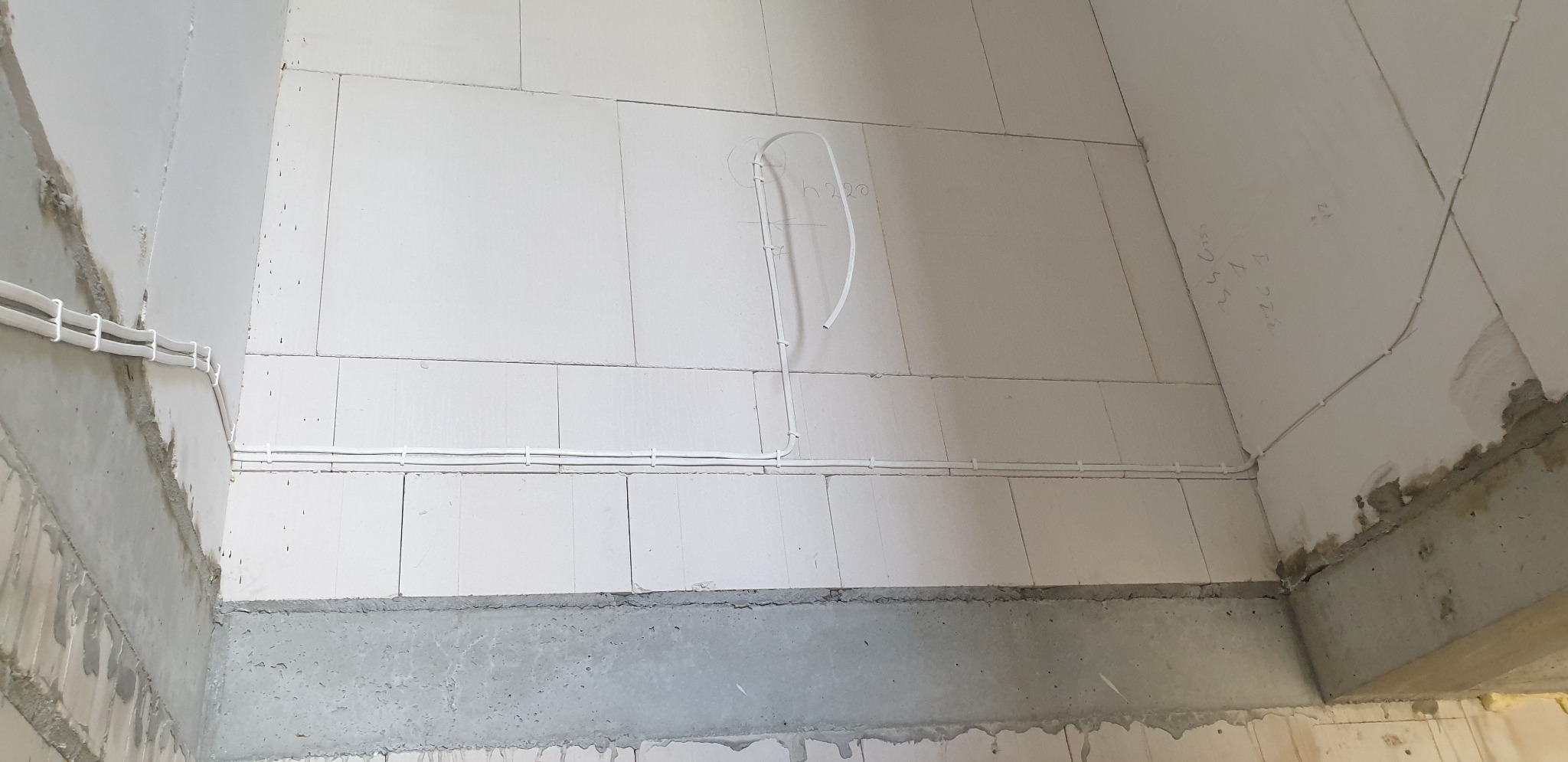 Surowy stan wnętrza z widocznymi przewodami elektrycznymi poprowadzonymi po ścianach z betonu komórkowego, zabezpieczonymi uchwytami, z widocznymi oznaczeniami na ścianie.